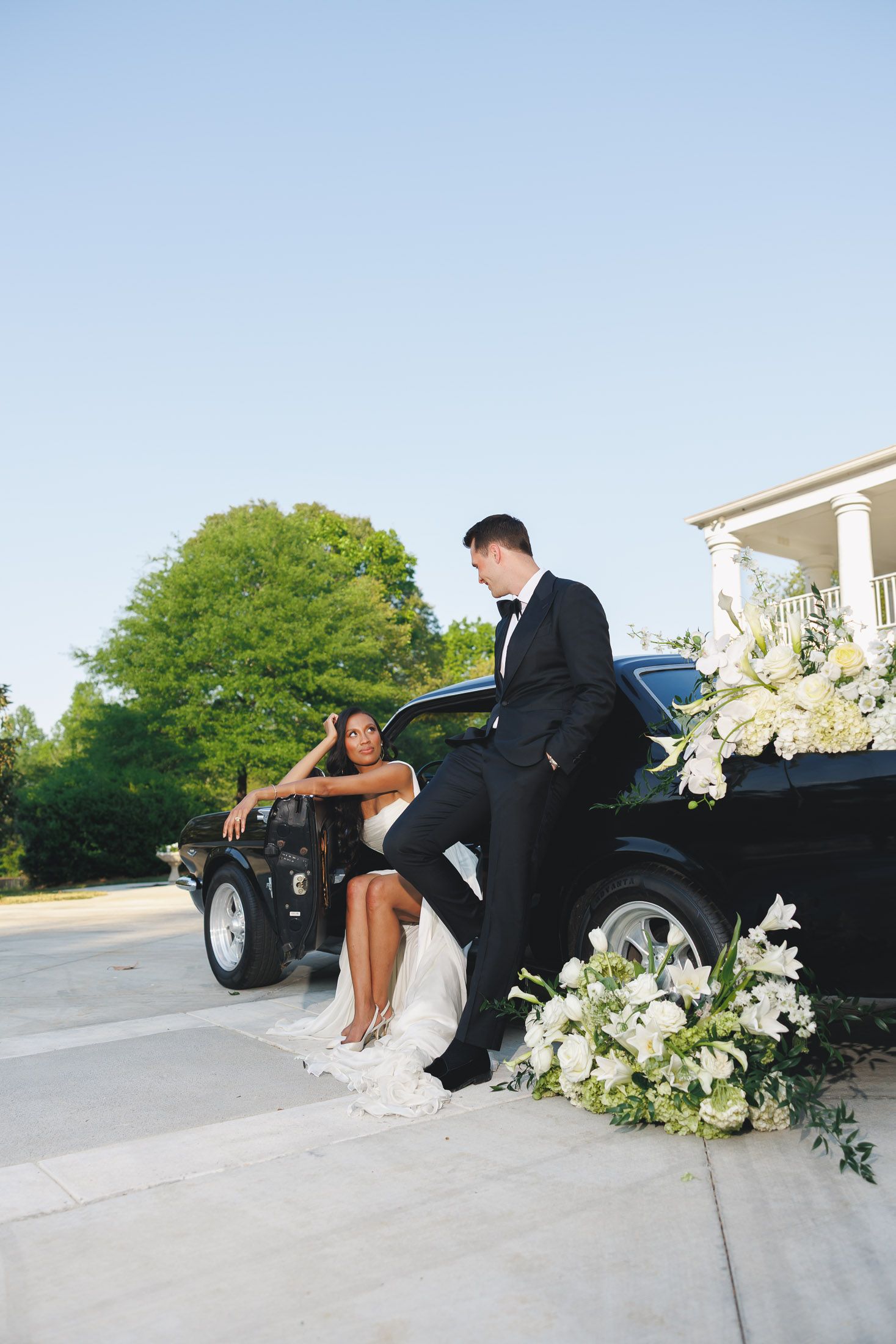 Bride and groom posing elegantly beside a black classic car adorned with white floral arrangements on their wedding day.