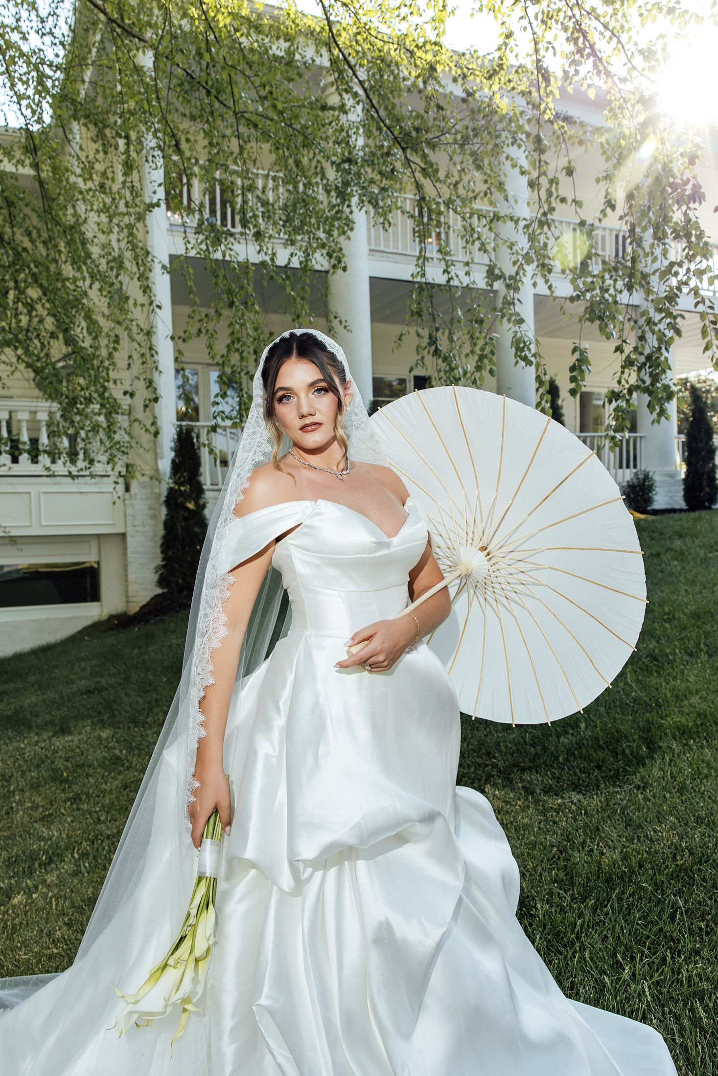 Bride in white off-shoulder gown with veil holds bouquet and parasol outdoors near elegant white mansion.