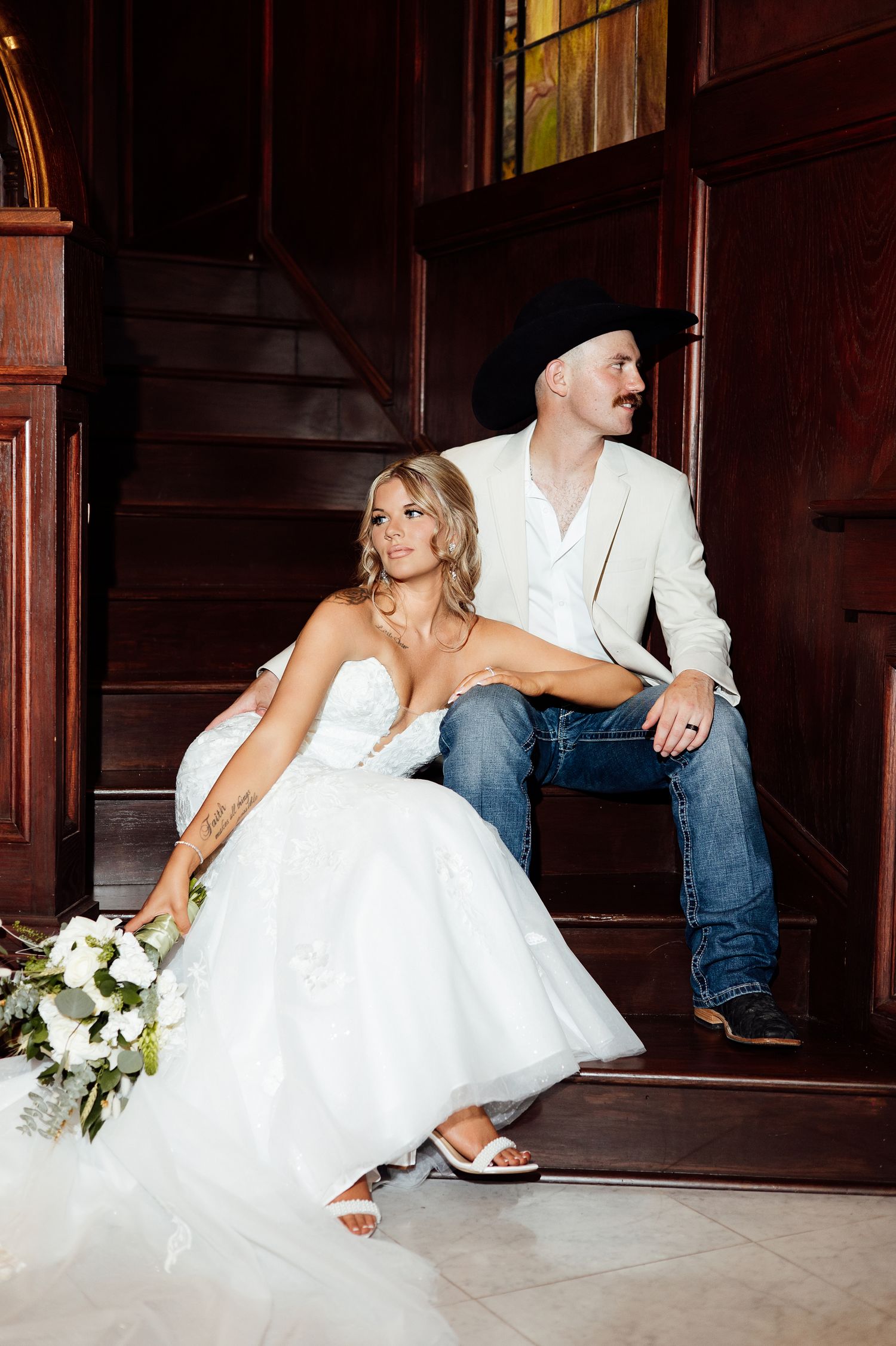 Bride in white strapless gown and groom in white blazer, jeans, and black cowboy hat sitting on wooden stairs.