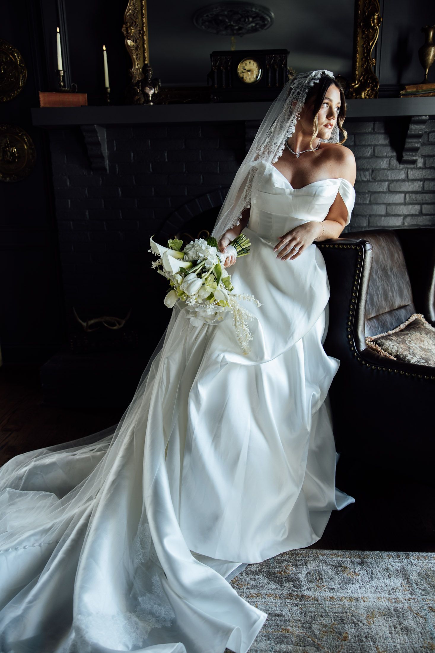 Elegant bride in off-shoulder white gown with veil holds white floral bouquet beside dark fireplace.