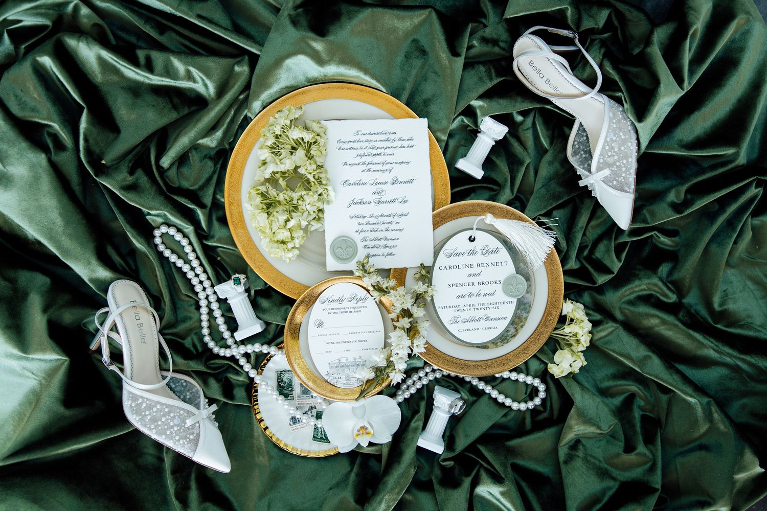 Elegant wedding stationery flat lay with gold-rimmed plates, pearl jewelry, and lace heels on green satin fabric.