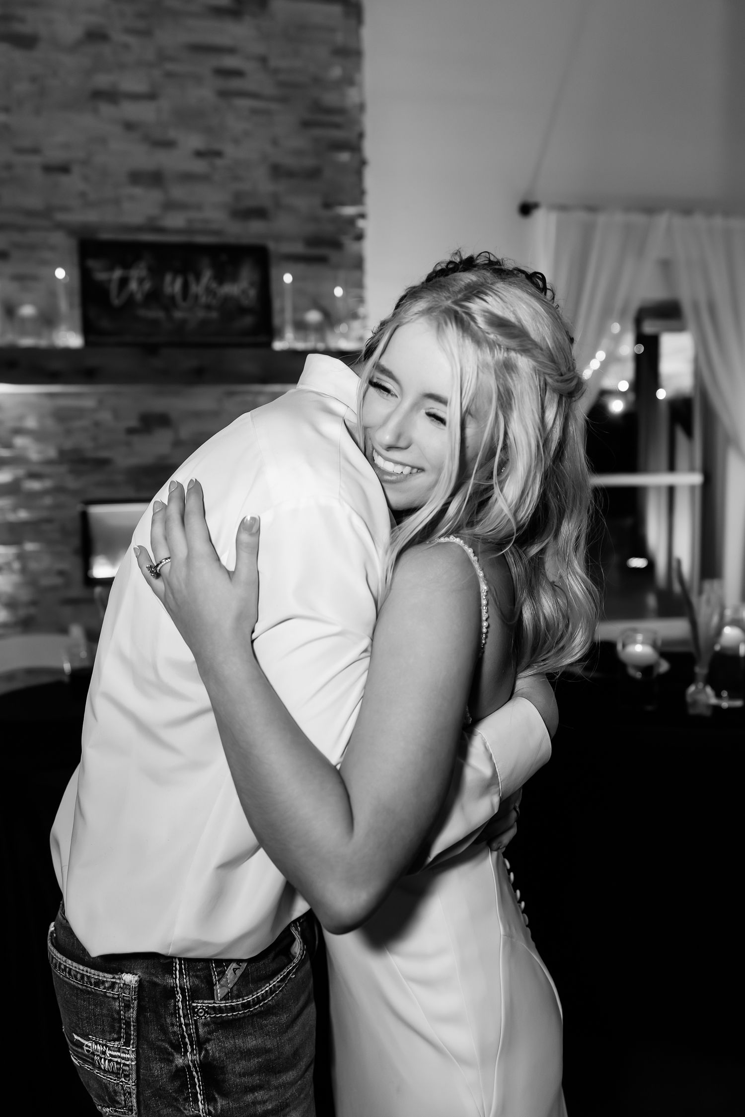 Black and white photo of a smiling blonde woman hugging a man in a white shirt at a wedding reception.