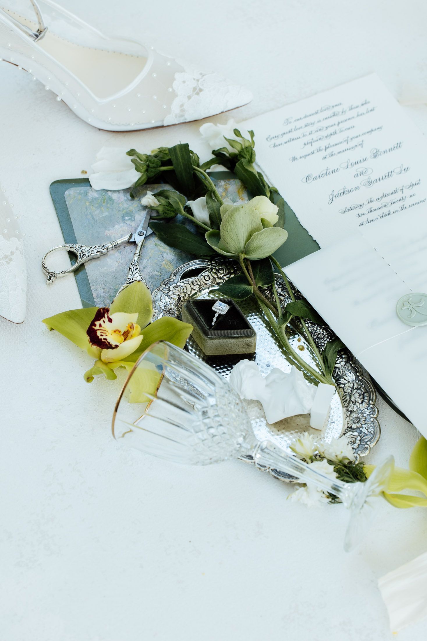 Wedding flat lay with lace heels, floral invitation, diamond ring box, crystal glass, and green orchids on white surface.