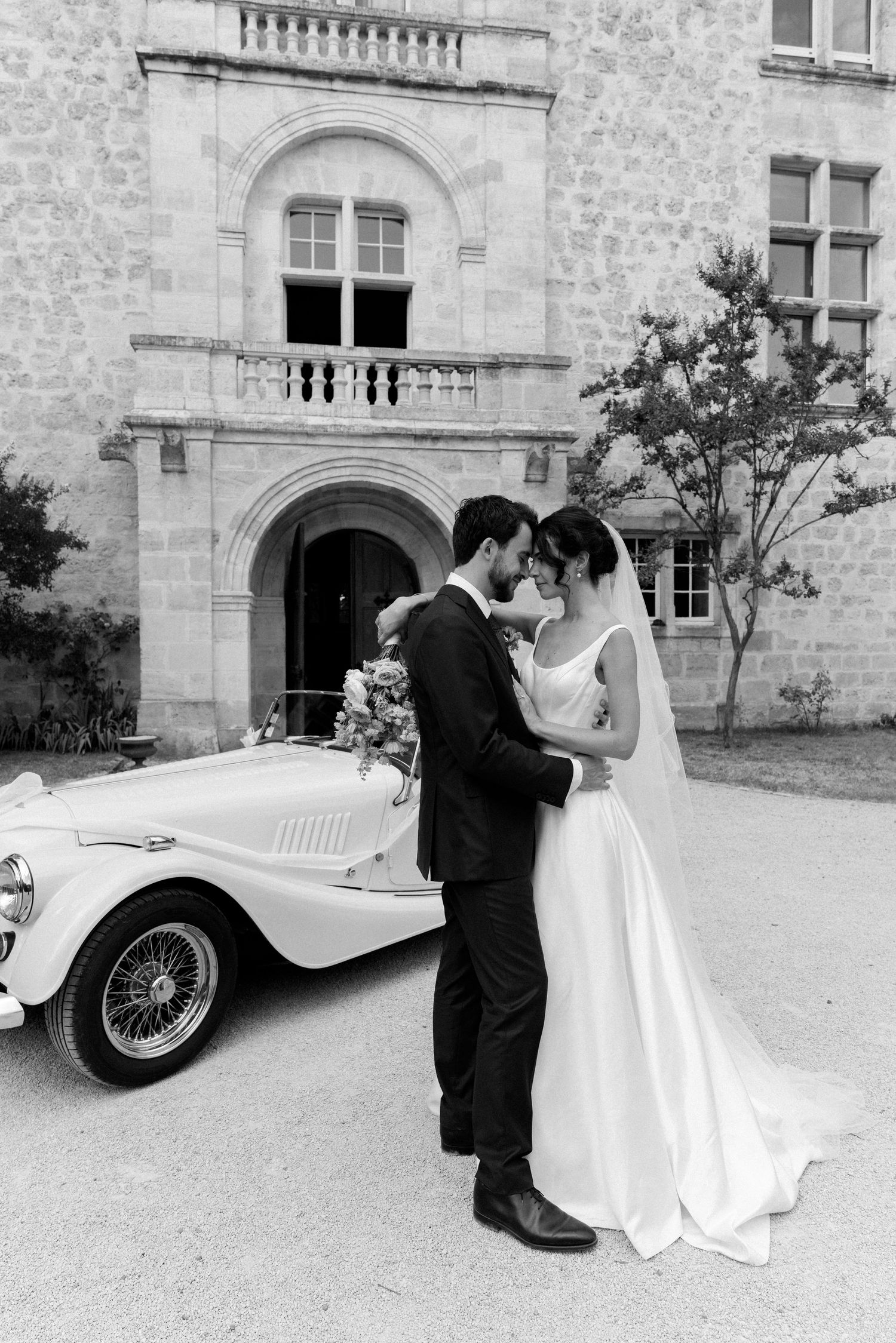 A classic white vintage car parked outside a stone building serves as backdrop for wedding photos.