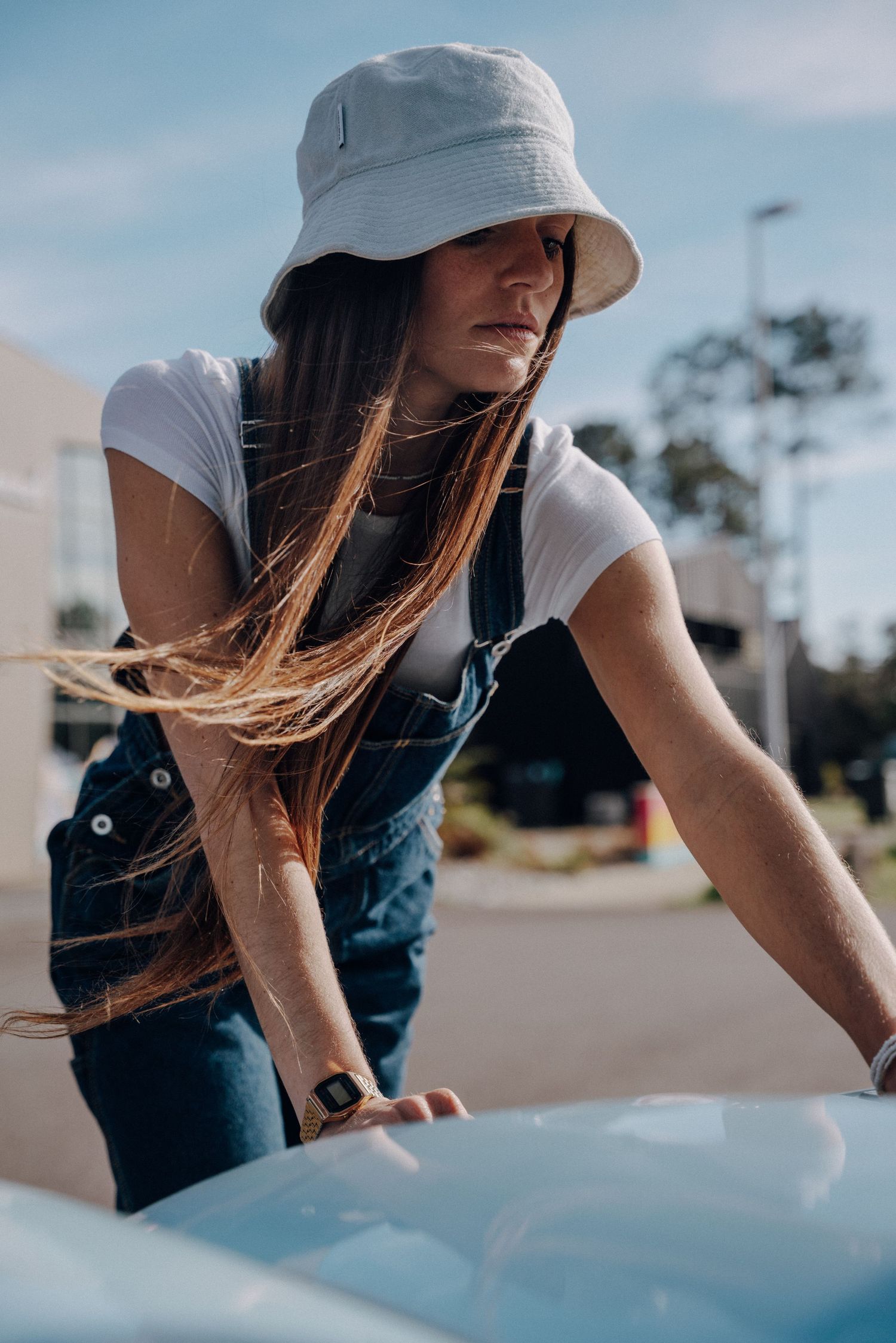 Person in denim overalls and sun hat leaning over light blue vintage car.