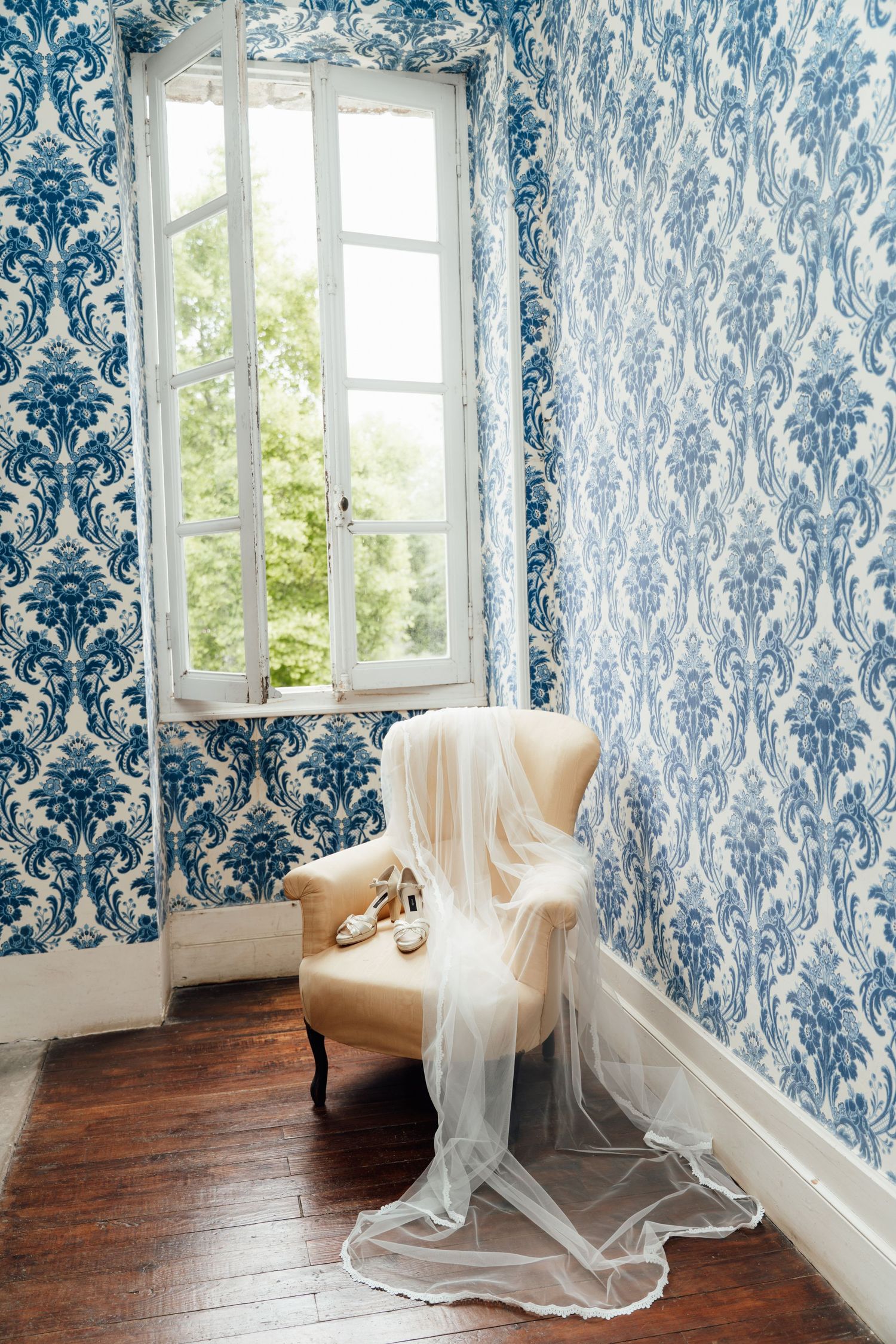 Elegant vintage armchair with bridal veil and wedding shoes in a room with blue damask wallpaper and white French window.