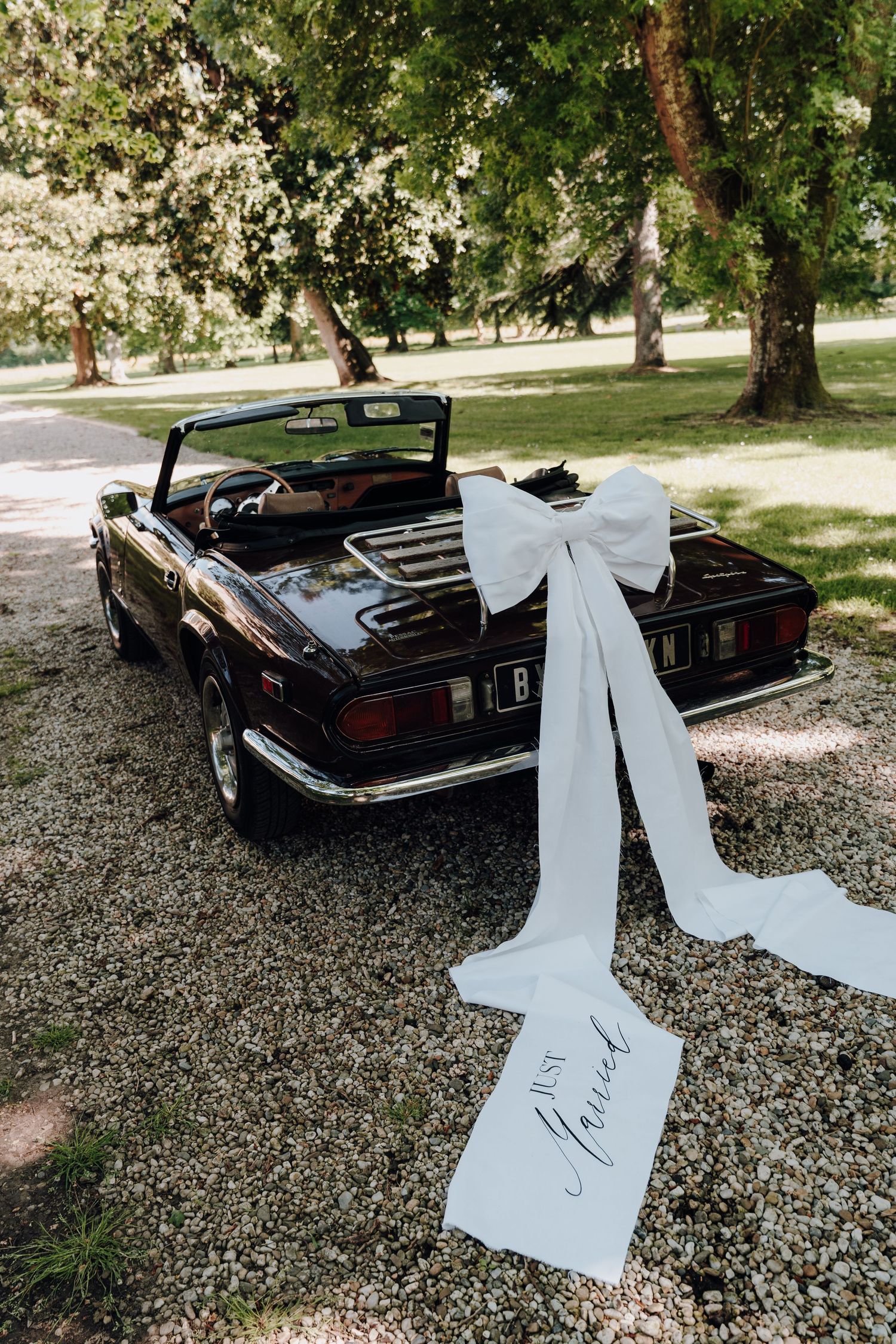 Classic black vintage car decorated with white wedding ribbons parked on gravel.