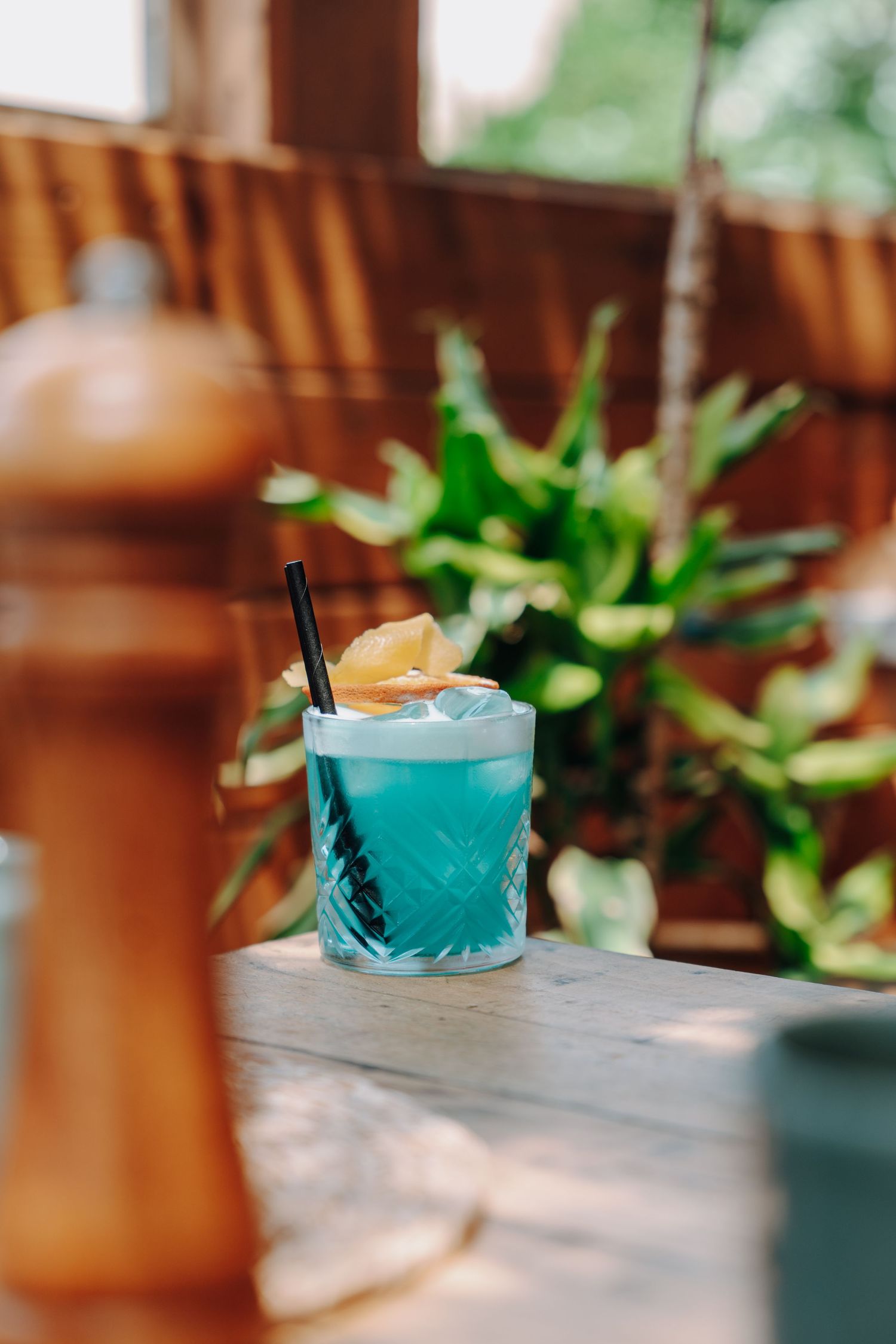A bright blue cocktail with foam topping and black straw sits on a wooden table in warm lighting.
