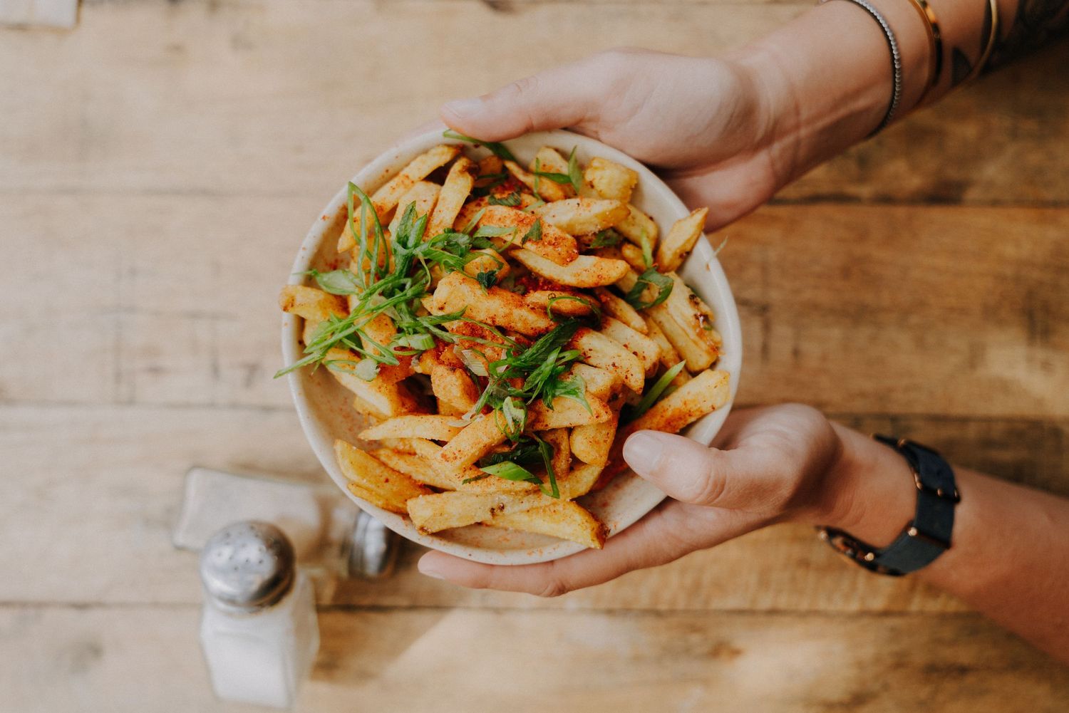 Hands holding a bowl of crispy french fries garnished with fresh herbs on a wooden table.
