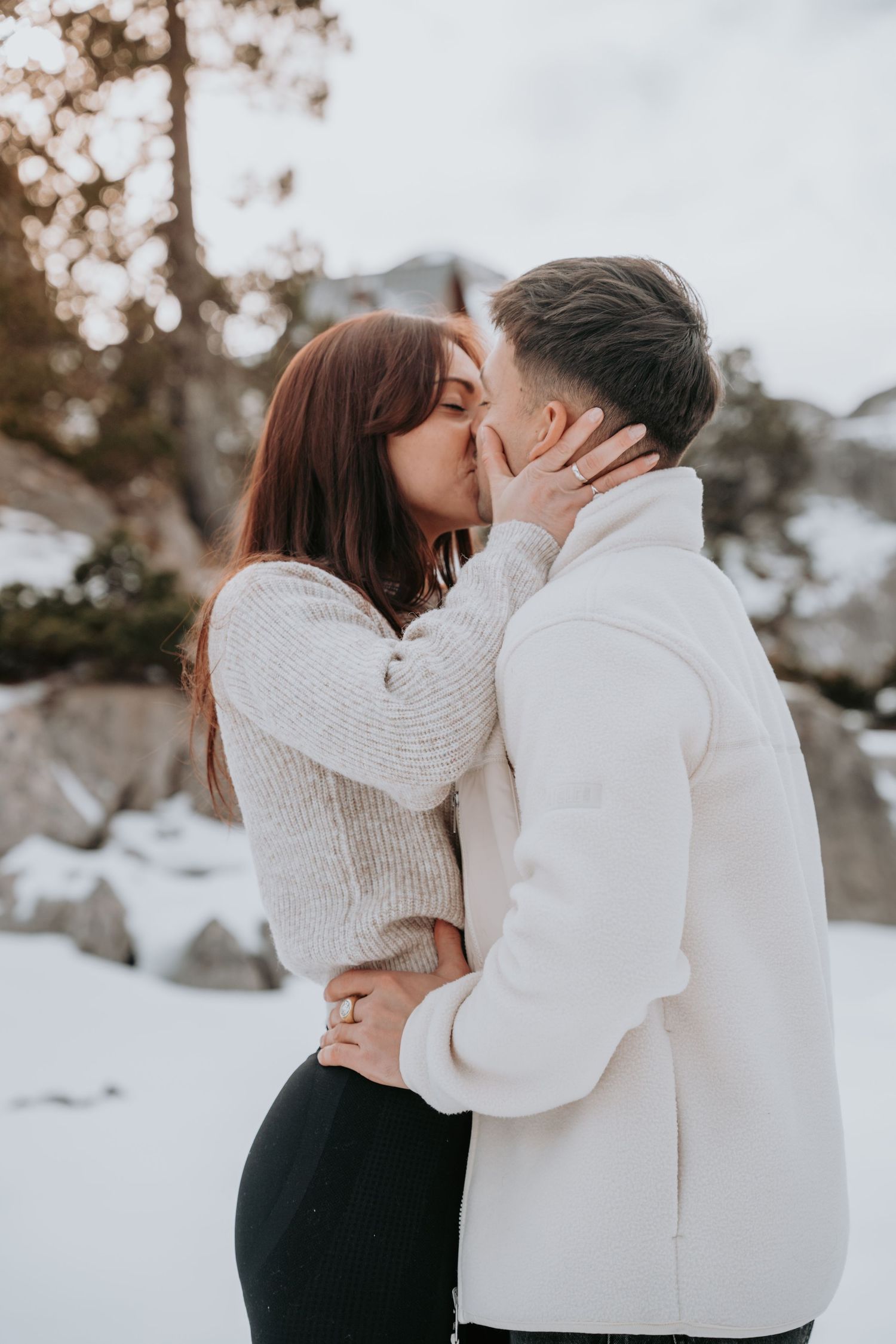 A couple shares an intimate moment in a snowy winter setting wearing matching light colored sweaters.