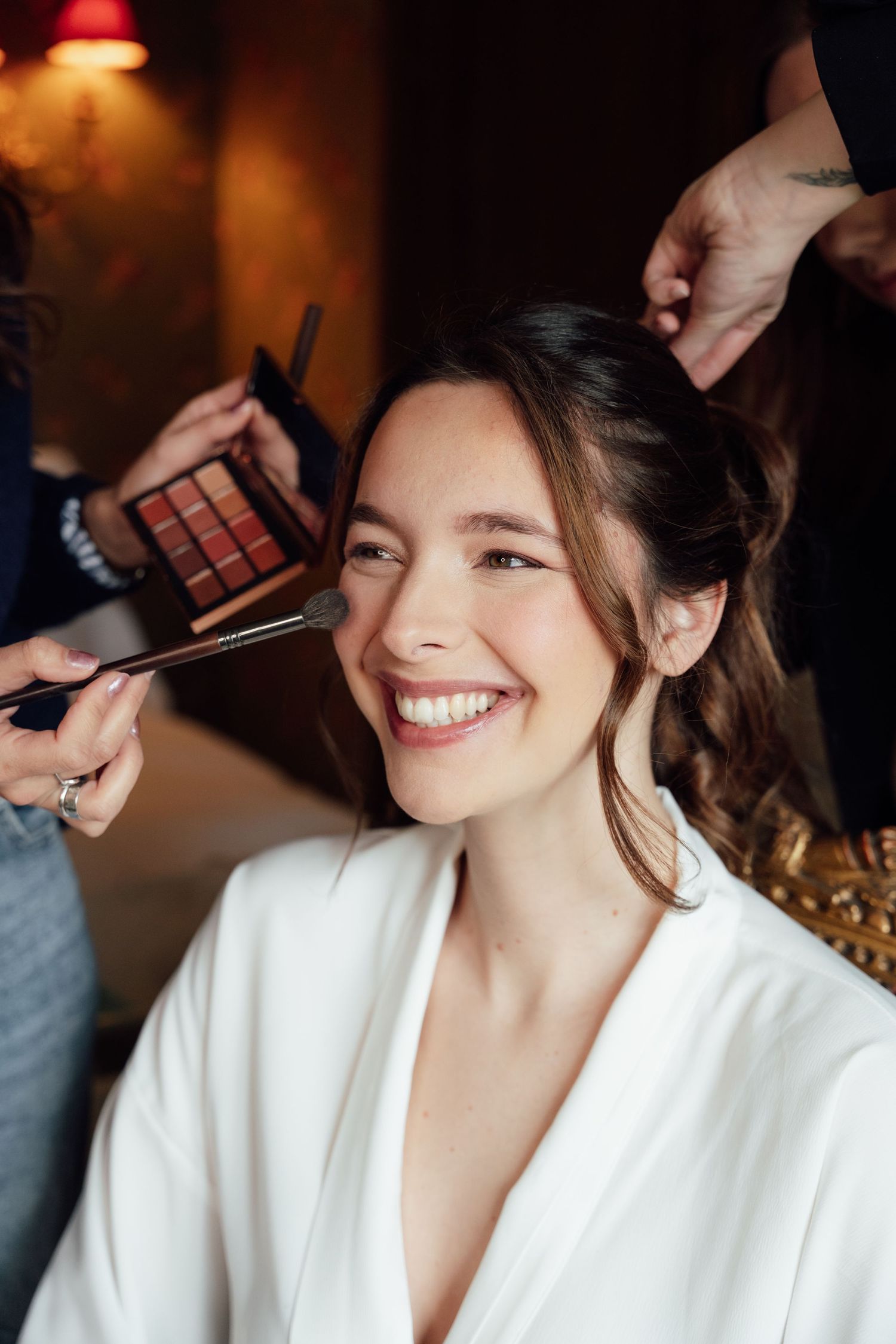 Smiling bride in white robe getting makeup applied with brush while hairstylist works on her hair.