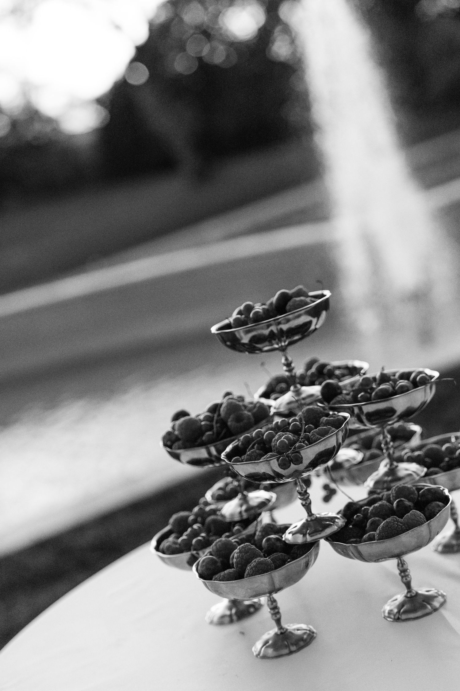 Black and white photo of elegant silver tiered dessert stand filled with fresh berries at an outdoor event.