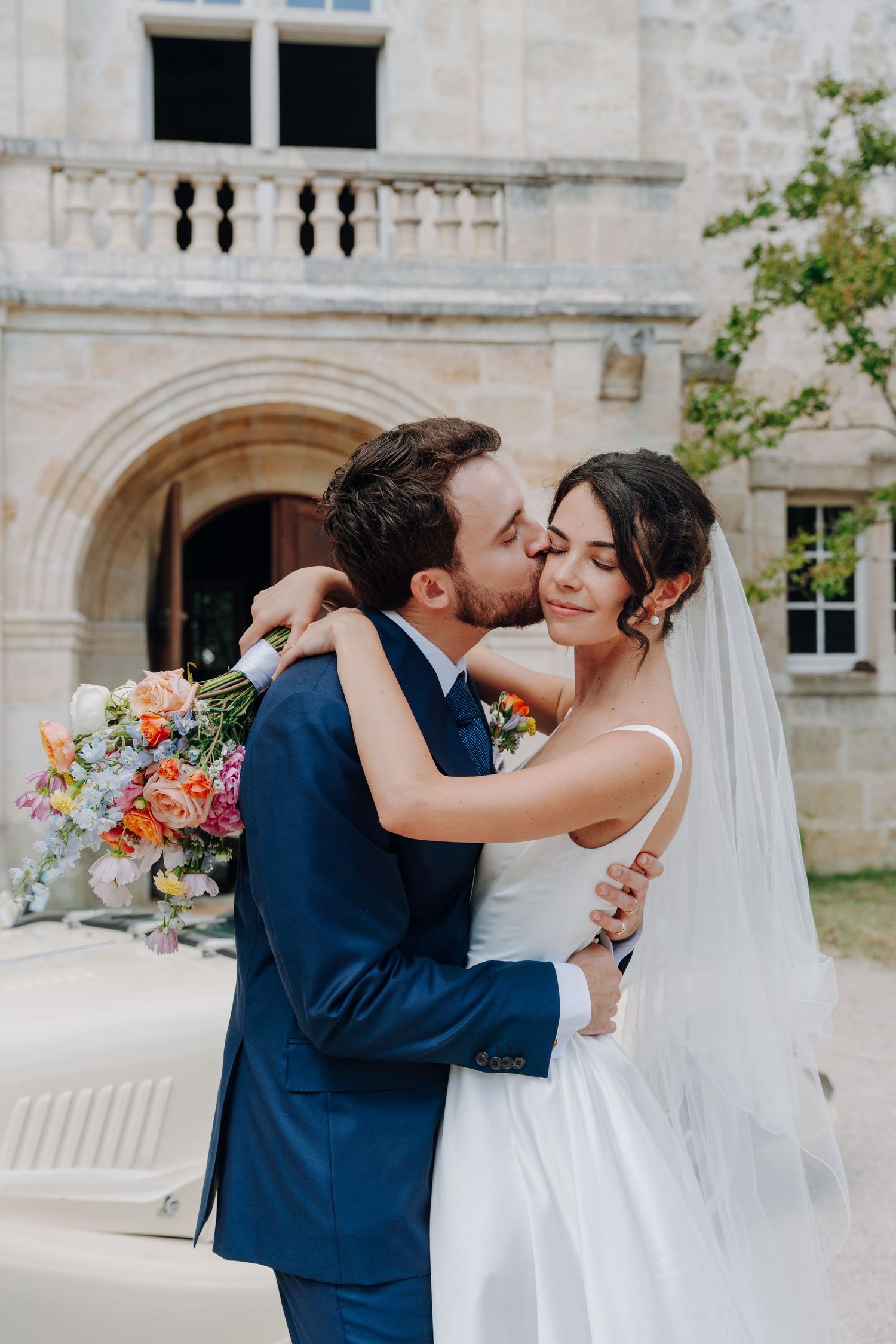 Newlyweds share romantic moment outside historic stone building with colorful bouquet.