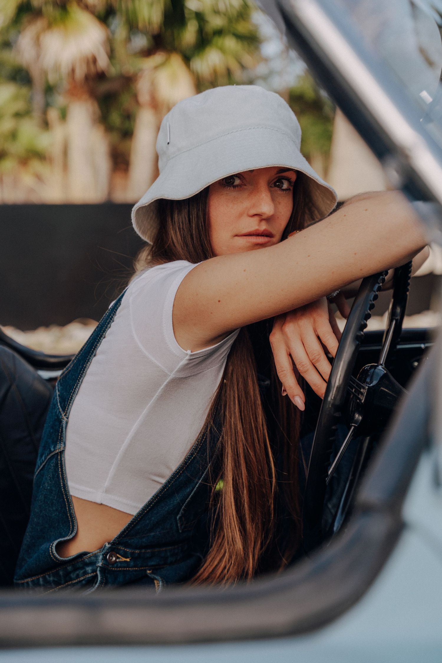 Relaxed poses through car window featuring person in white shirt and sun hat with vintage vehicle.