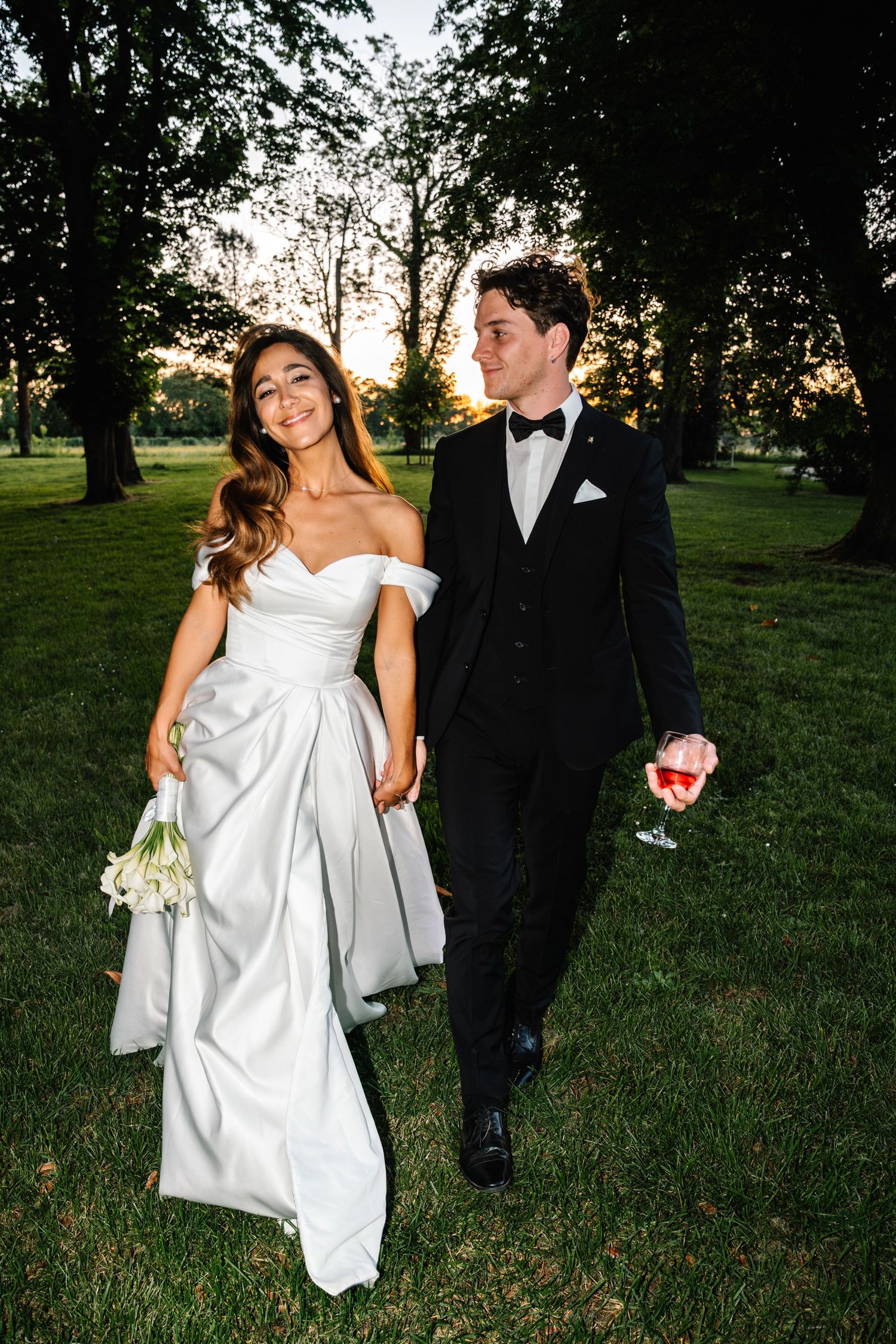 A couple walks through a grassy park at sunset, with the bride wearing a flowing white gown.