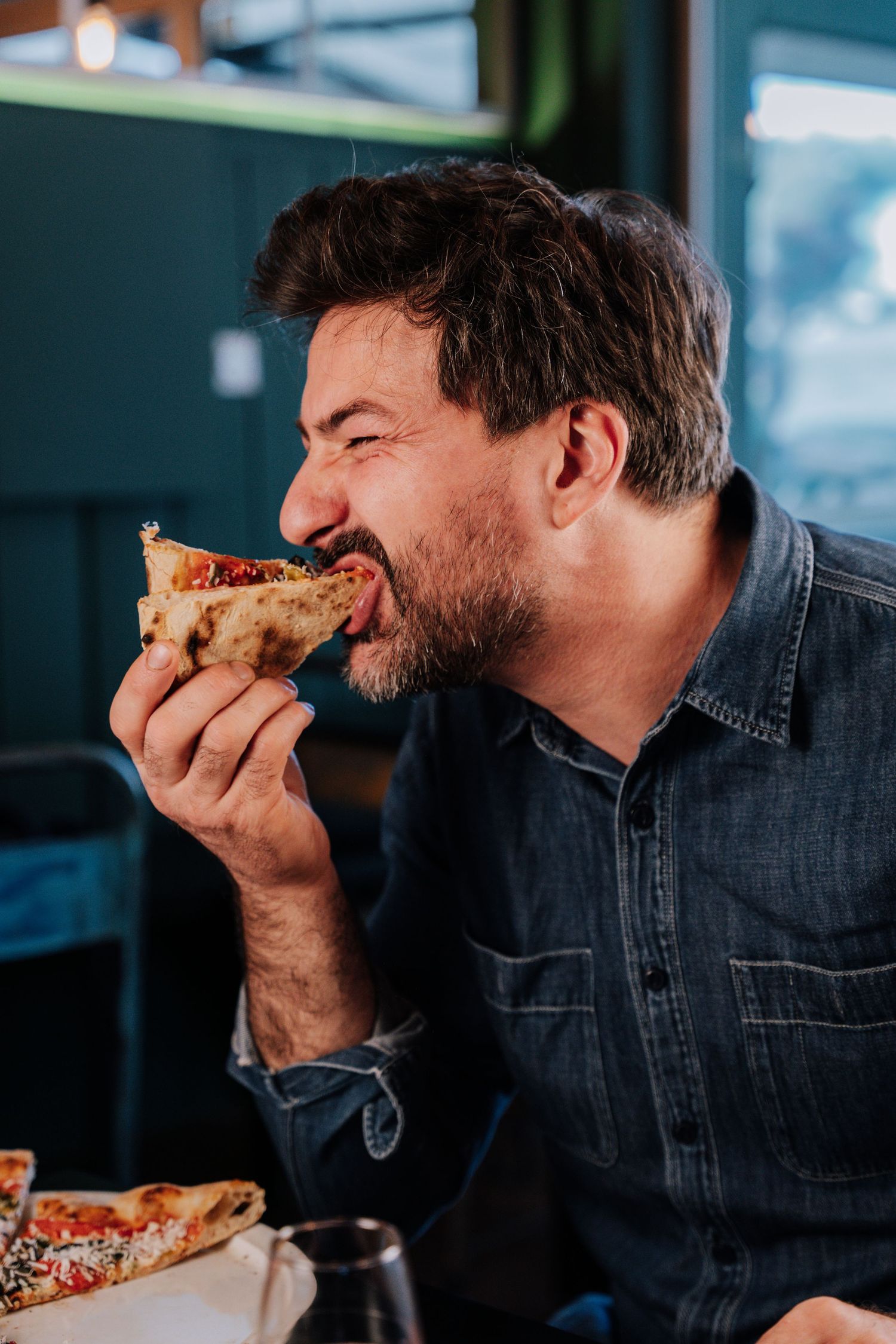 A person in a denim jacket takes a big bite of a slice of pizza in a sequence of progressive eating shots.
