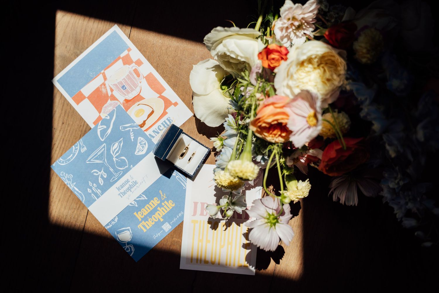 Wedding flat lay with colorful bridal bouquet, ring box, and blue stationery on dark surface in natural light.
