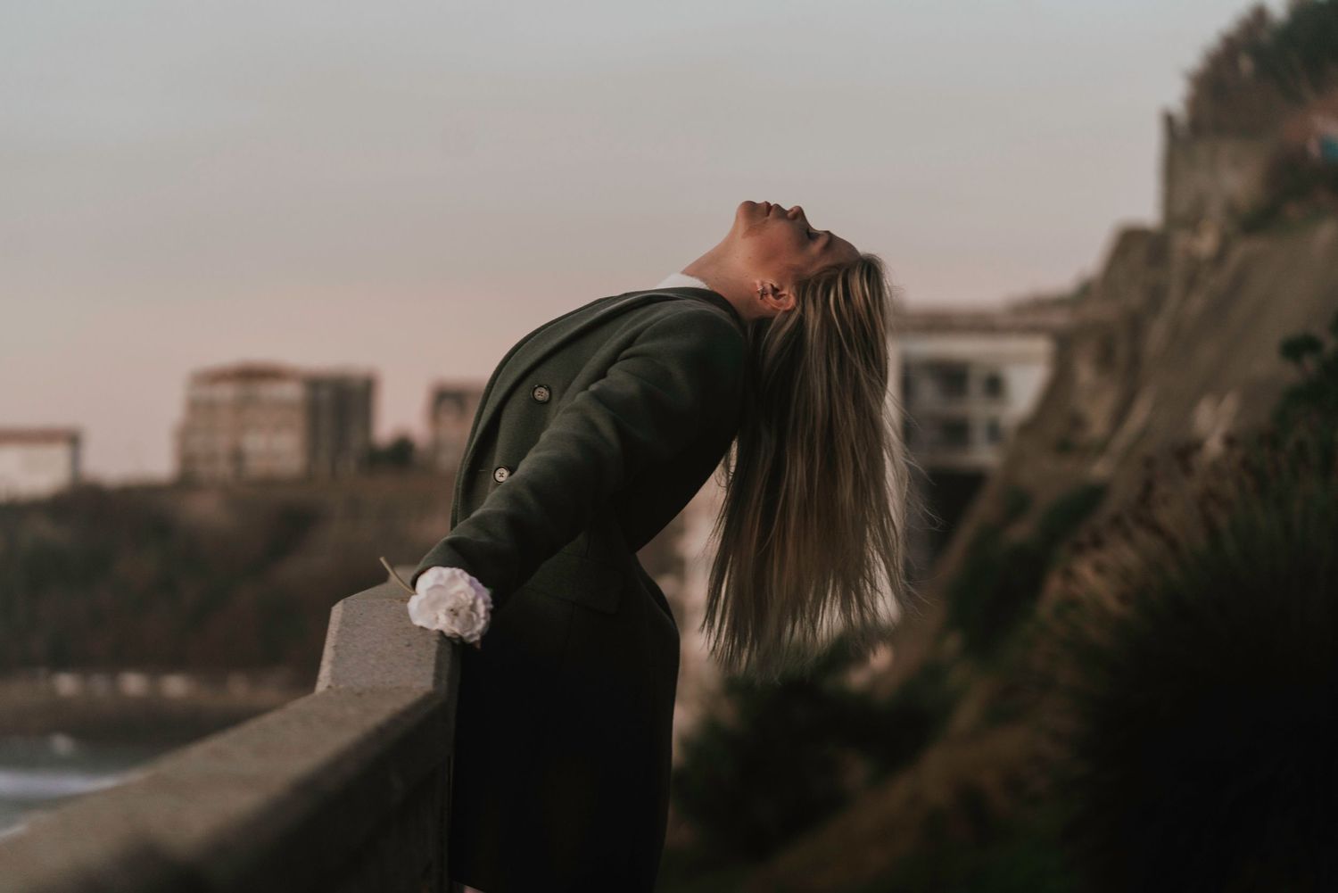 A person in a dark jacket leans back against a railing at sunset with an urban backdrop.