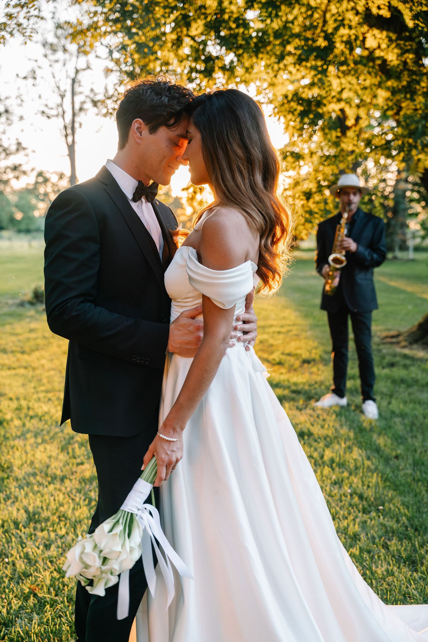 Romantic sunset wedding photo with couple embracing while musician plays in background.