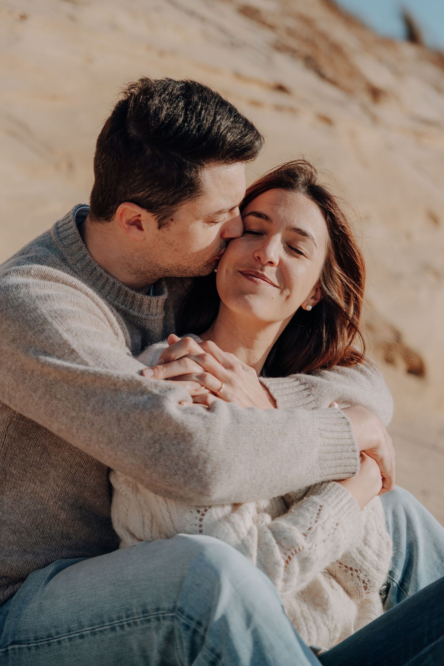 A romantic couple embraces on a beach while wearing cozy sweaters during a tender moment.