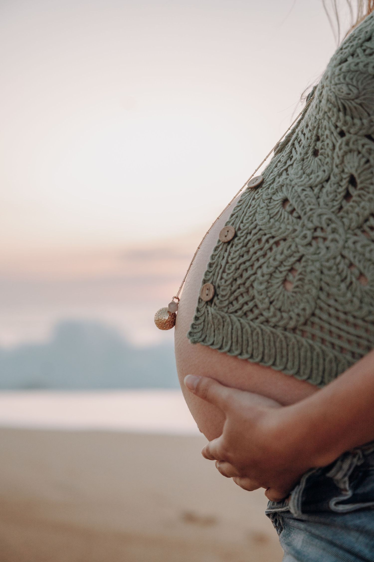 Close-up details of pregnant belly wrapped in knit sweater against beach backdrop.