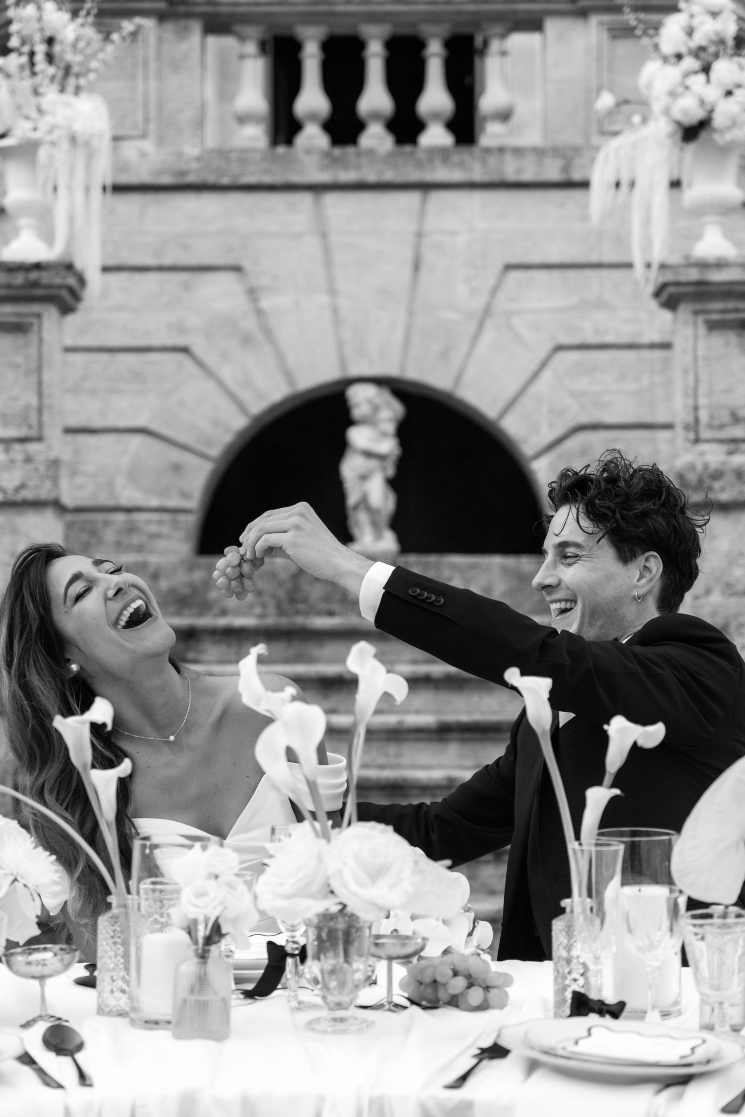 Black and white wedding photo of laughing bride and groom sharing grapes at elegant outdoor reception table.