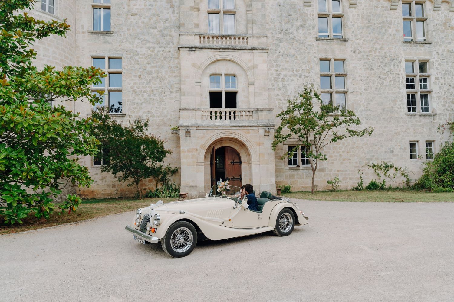 Vintage cream-colored classic car parked in front of a historic stone castle building.