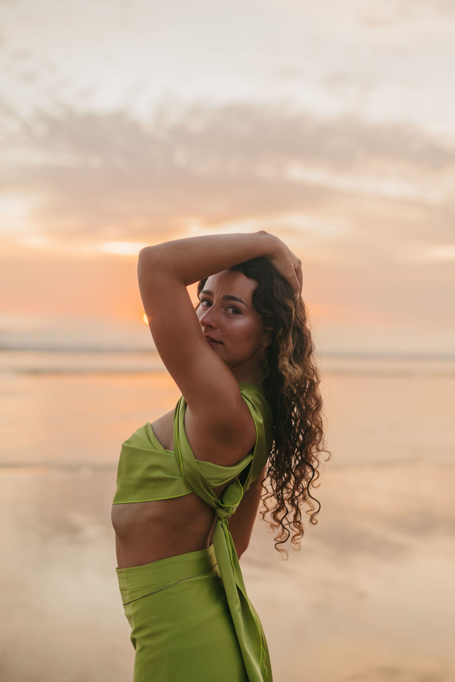 A woman in a lime green outfit poses against a dreamy sunset sky with warm orange and pink hues.