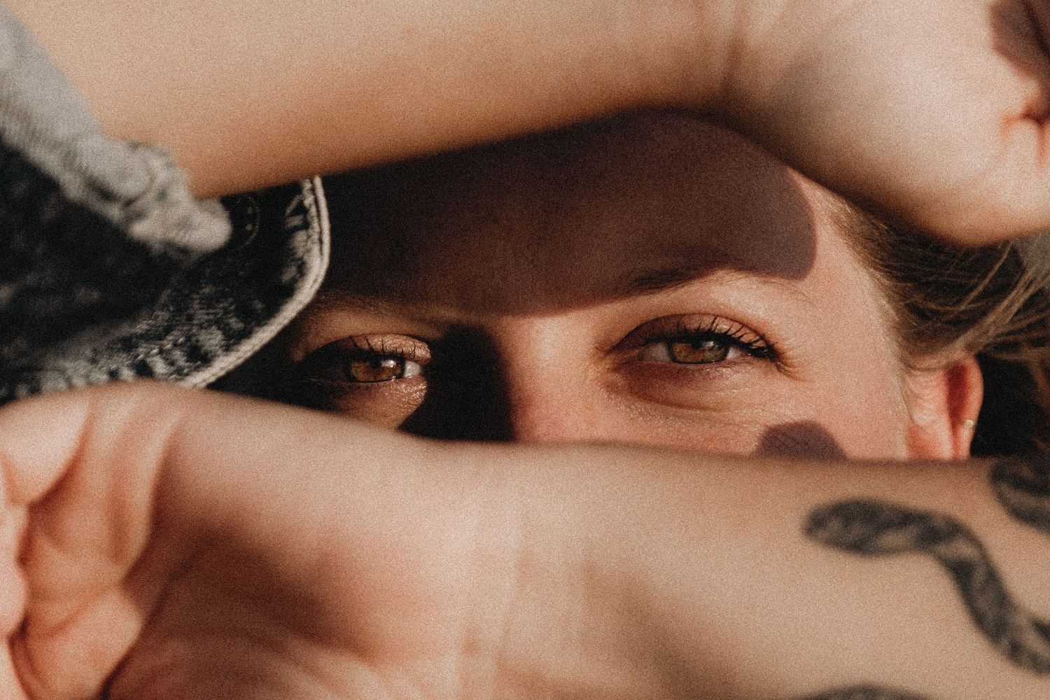 Close-up artistic photo of eyes peeking through shadows with dramatic lighting and tattoo visible.