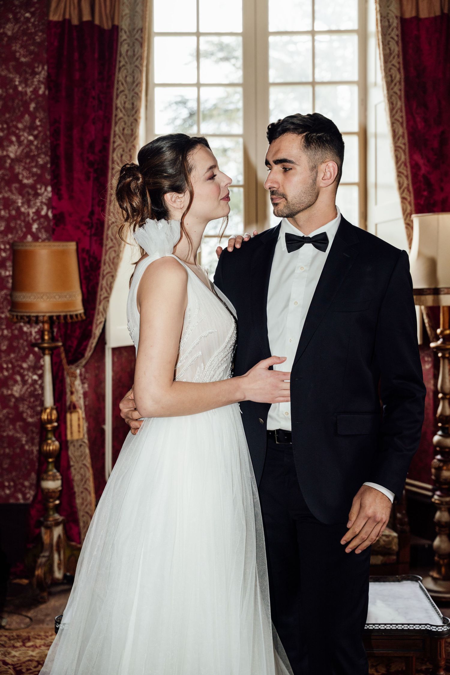 Elegant bride and groom embracing in a luxurious red-draped room, wearing white gown and black tuxedo.