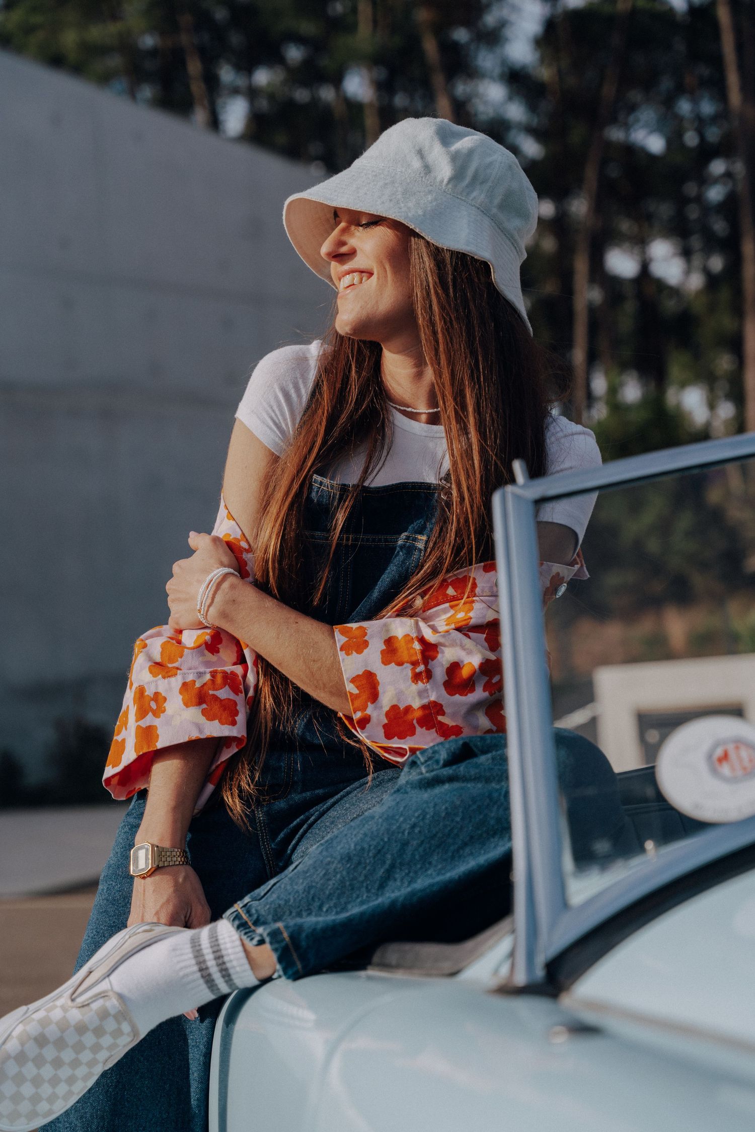 Woman in casual attire and white hat sitting on a vintage car with floral print details.
