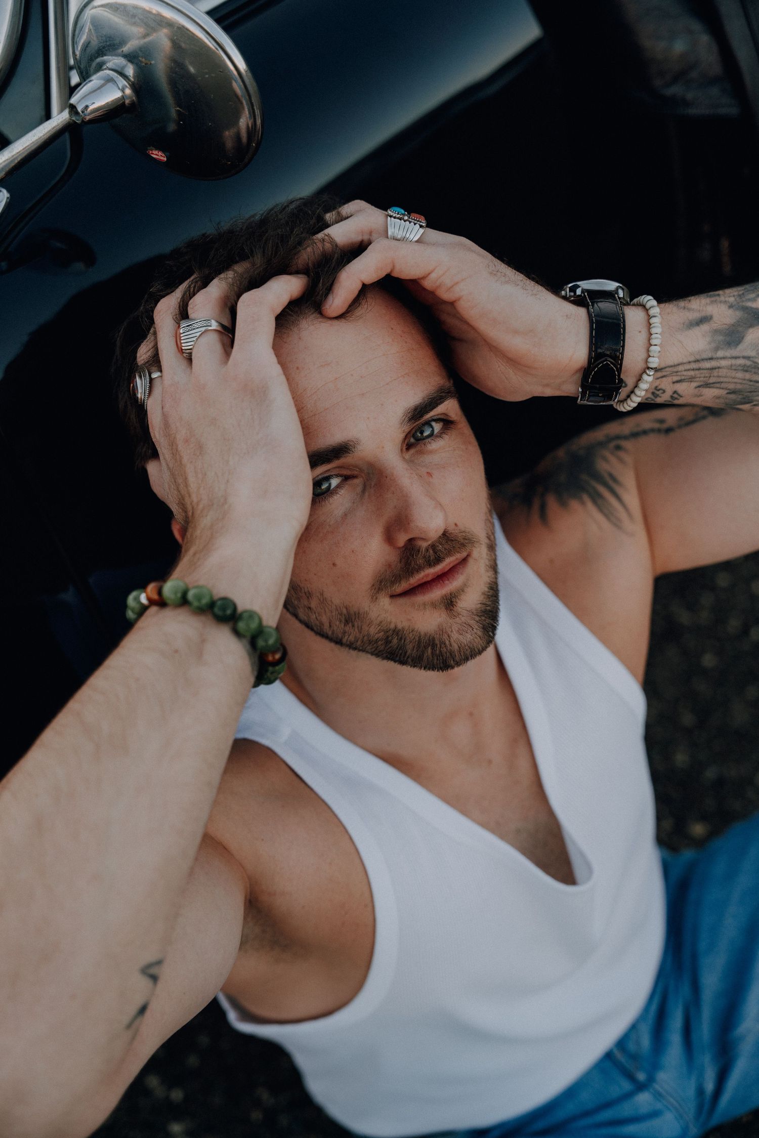 Close-up portrait in white tank top showing tattoos and jewelry against dark background.