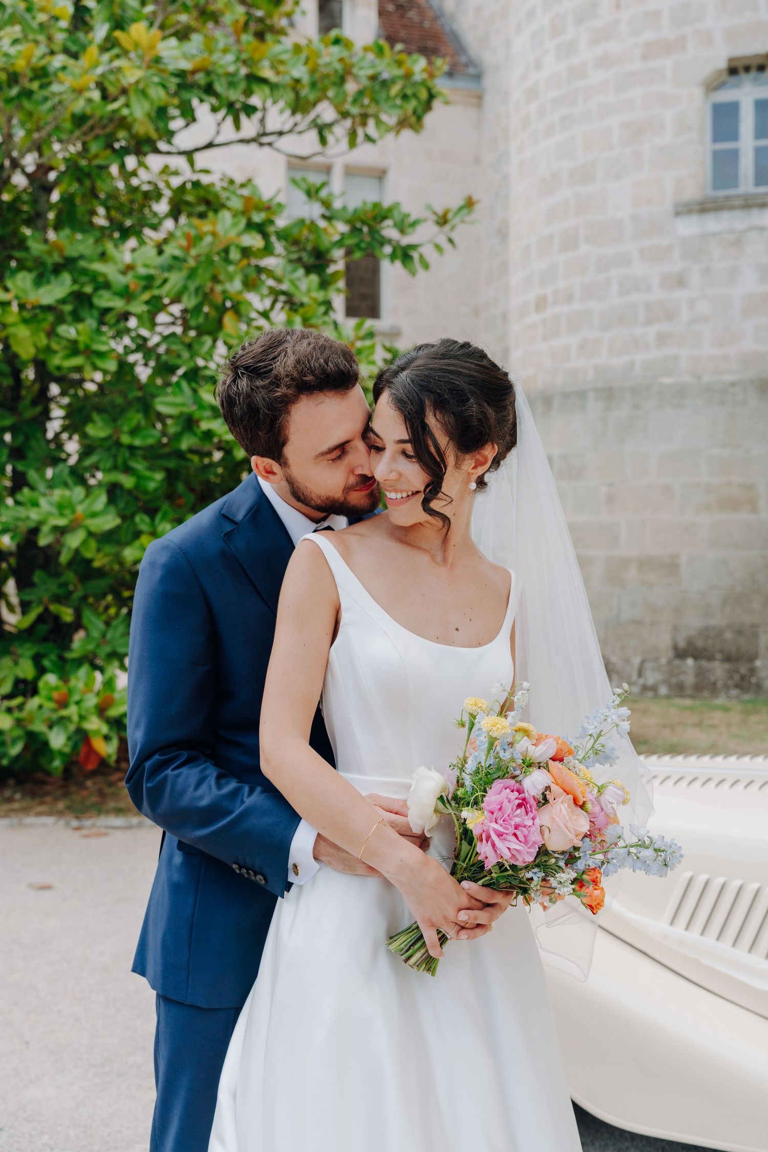 Bride and groom embrace near stone building surrounded by greenery and holding floral bouquet.
