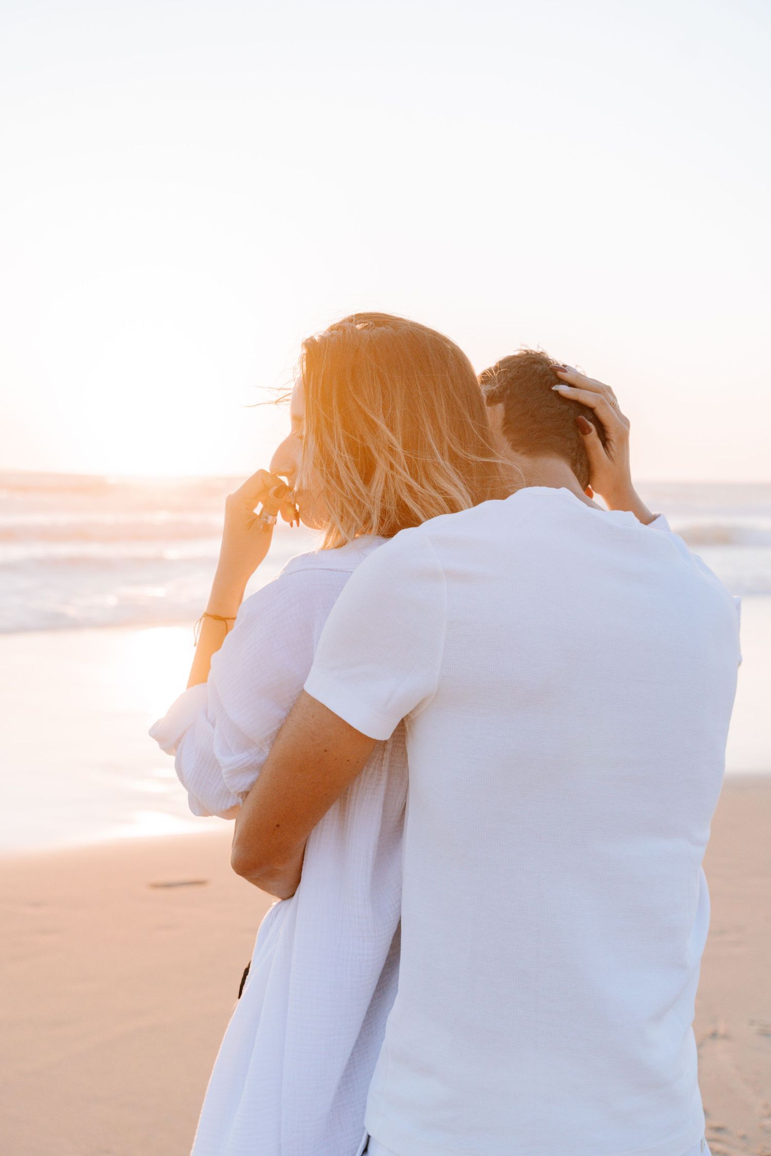 A couple in white clothing embraces while watching the sunset on a peaceful beach.