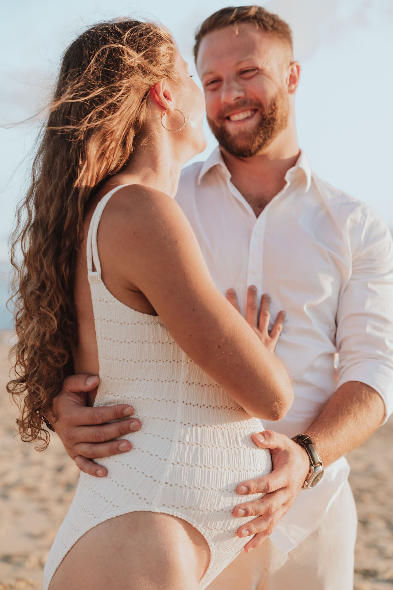 Couple in white outfits embracing on beach during maternity photoshoot.