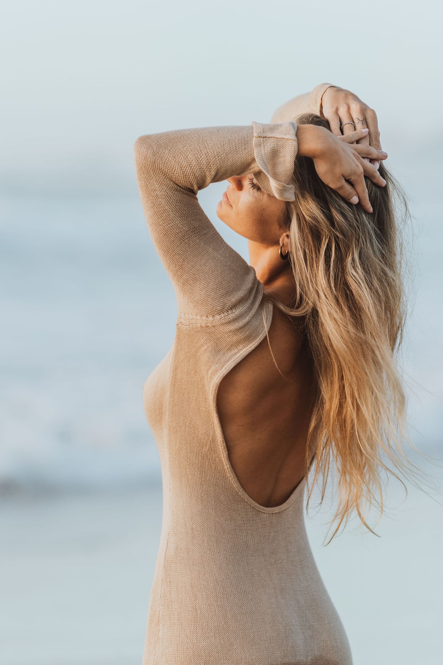 Woman in beige knit dress poses expressively against ocean backdrop.