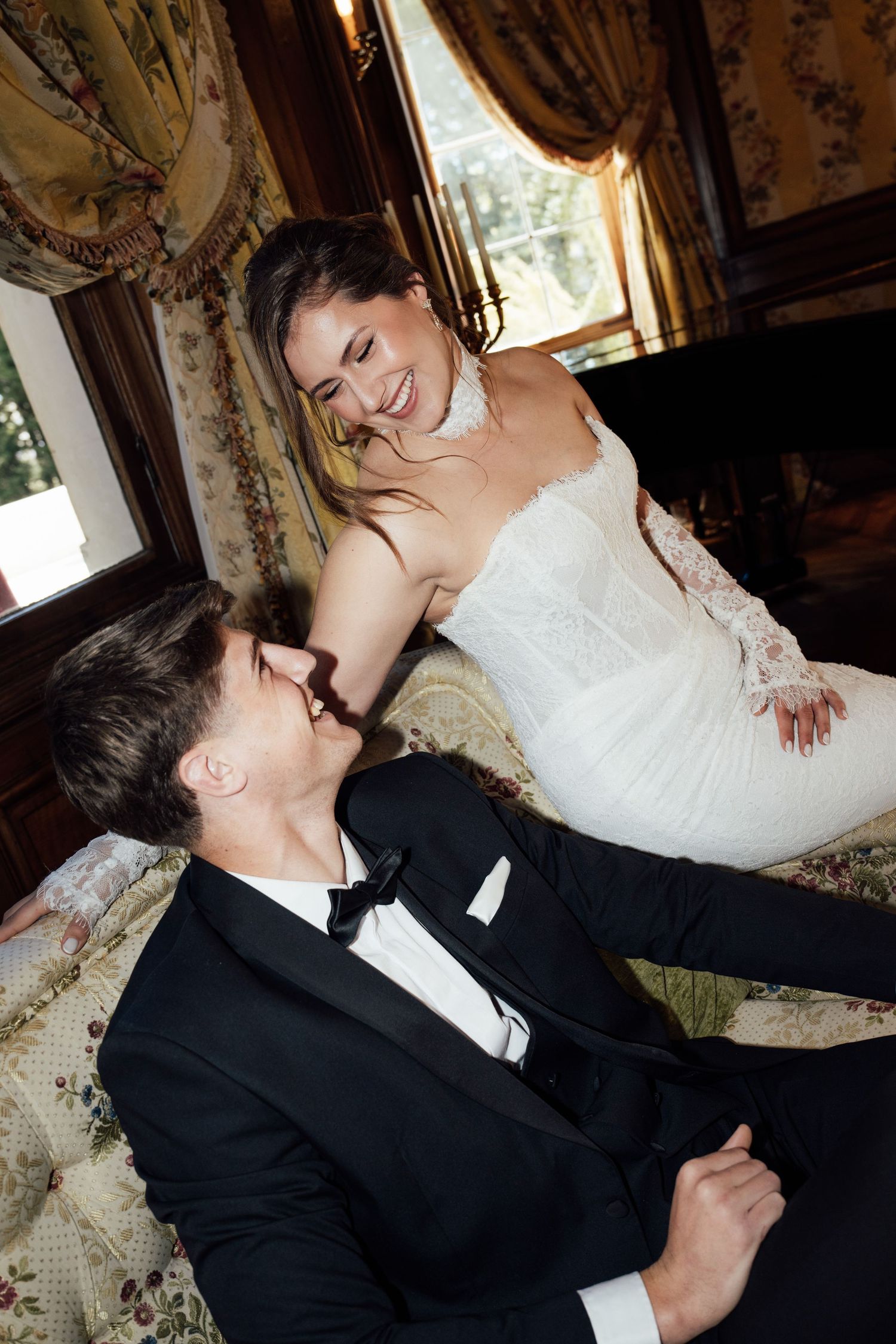 Bride in white lace gown and groom in black tuxedo share a joyful moment on a floral sofa in an elegant room.