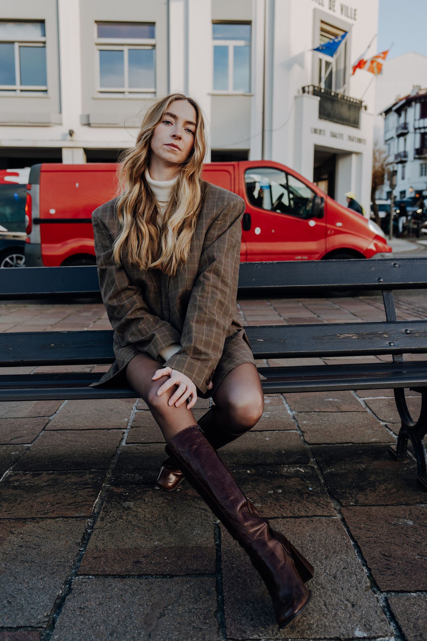 Person in plaid blazer and boots sitting on urban bench during golden hour.