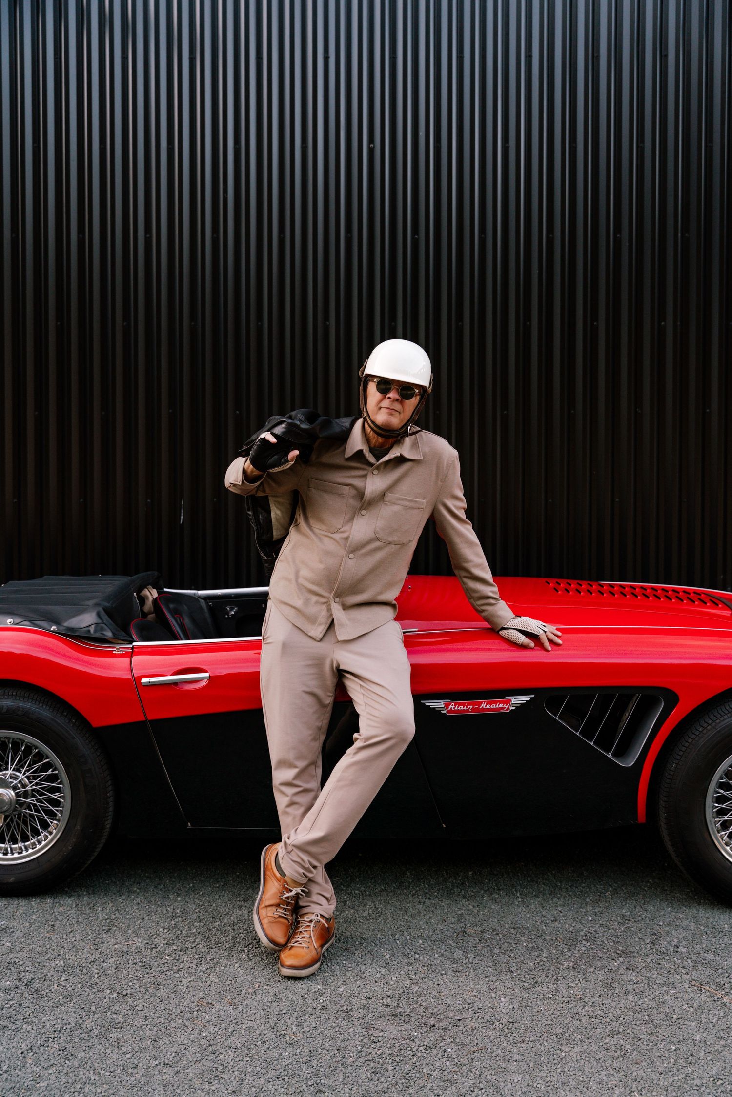 Person in beige jumpsuit poses playfully around a red vintage sports car against dark background.