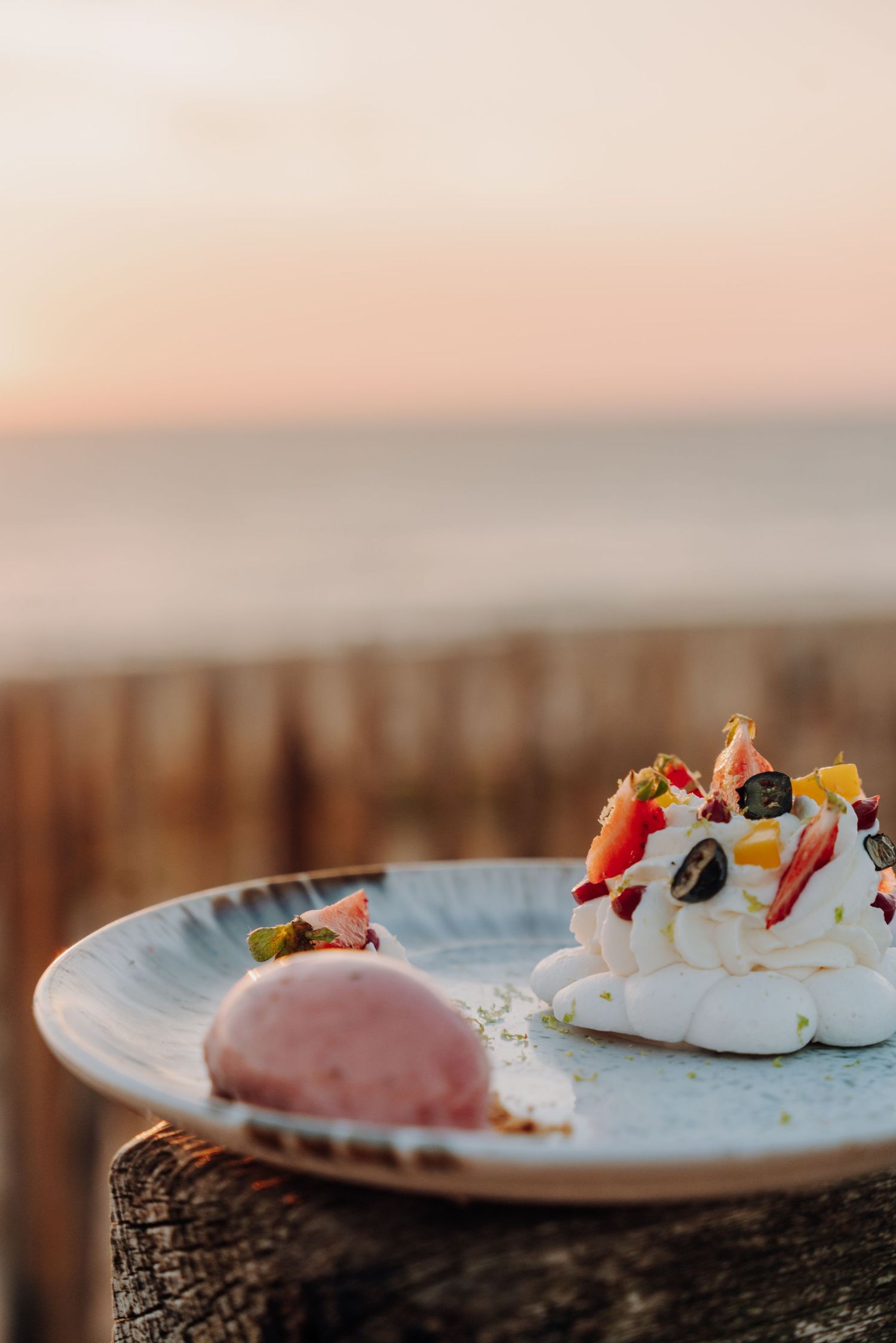 A row of Pavlova desserts with fresh fruit toppings sit on plates against a blurred sunset background.