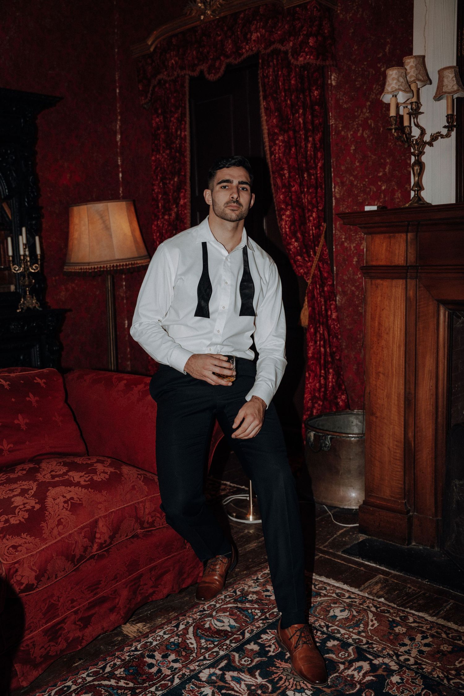 Man in formal attire relaxing on red velvet couch in vintage-styled room.