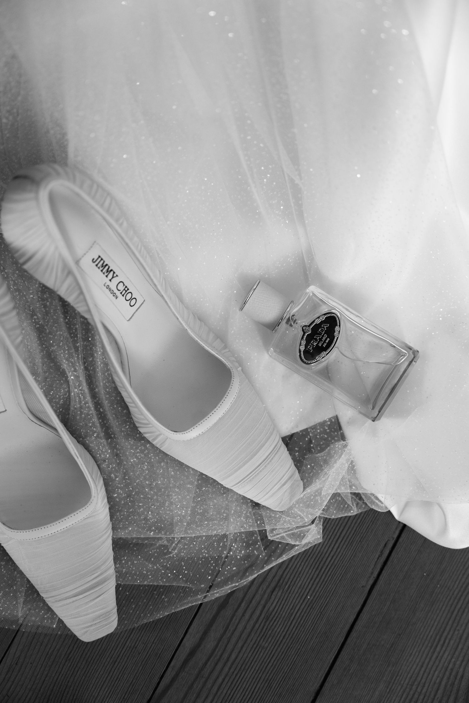 Black and white photo of Jimmy Choo bridal heels, perfume bottle, and tulle wedding dress on wooden floor.