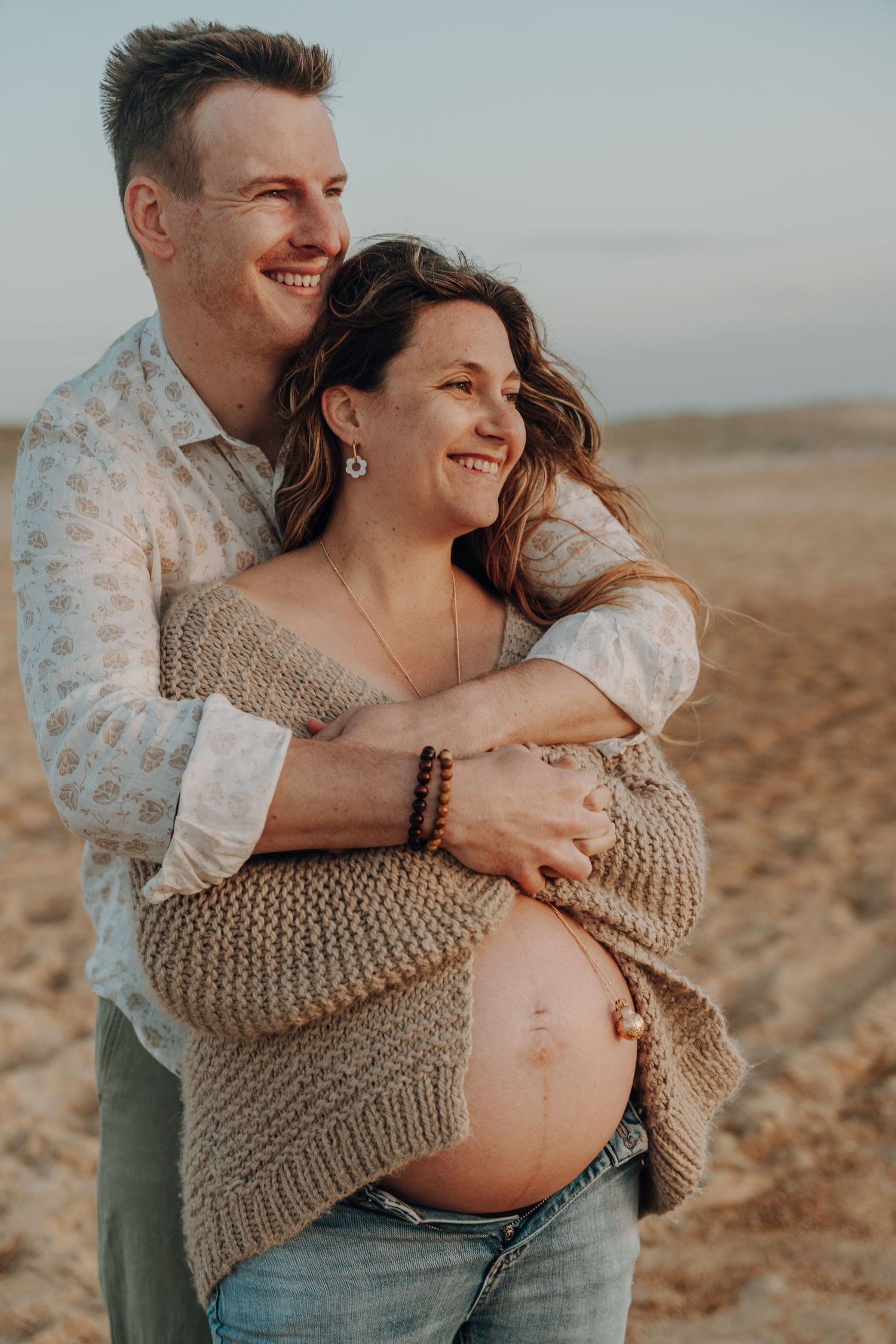 Maternity photos showing couple in casual wear embracing on sandy beach.