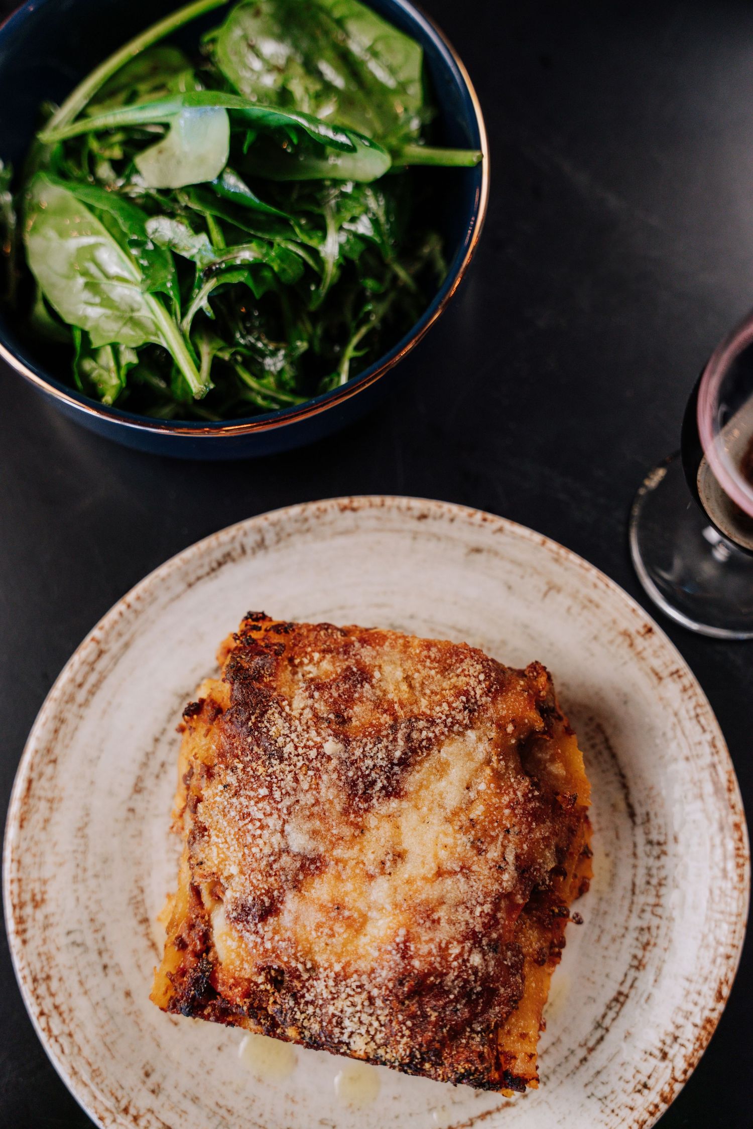 A rustic plate of lasagna served with a side salad of fresh greens and a glass of red wine on a dark table.