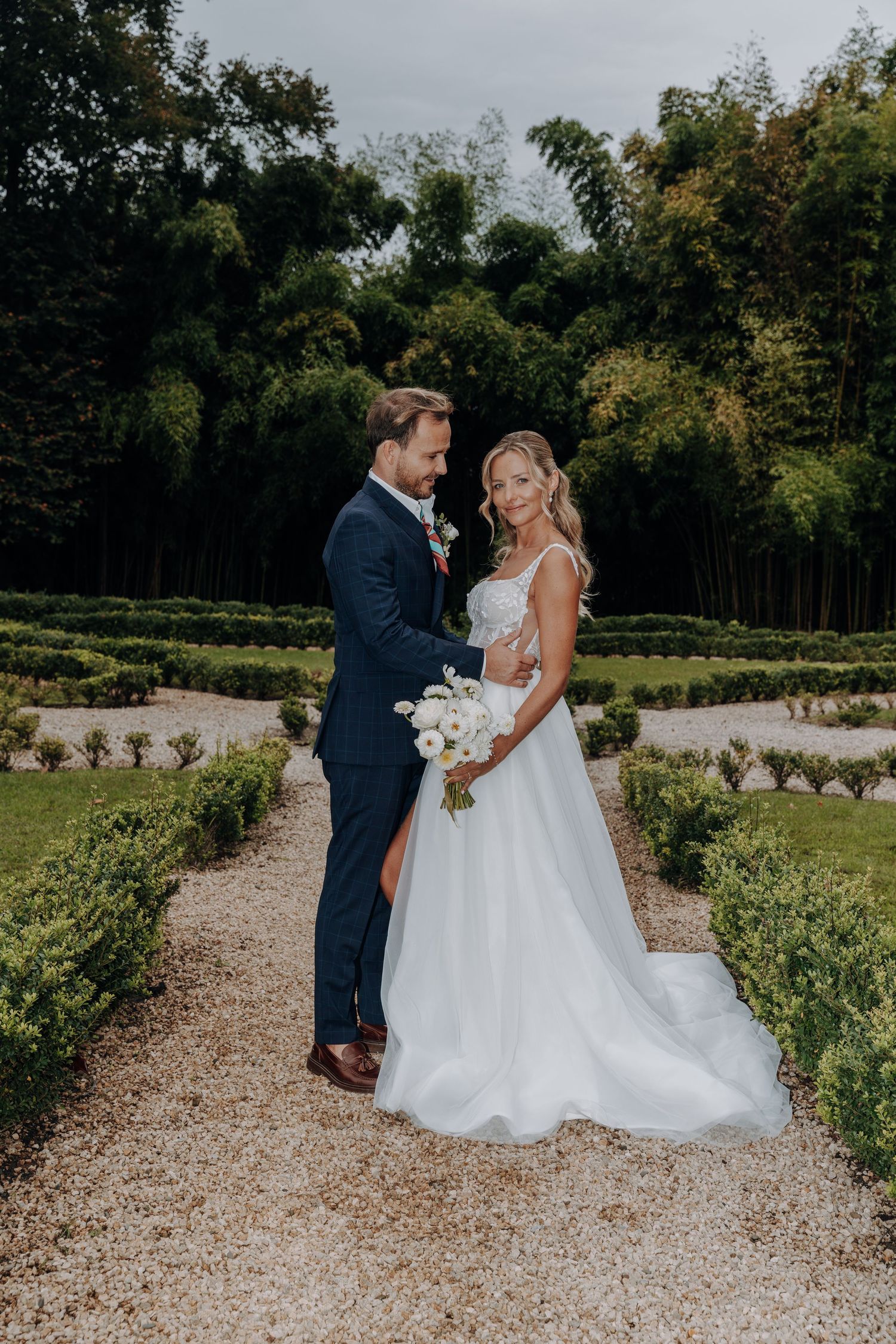 Newlyweds stand together on garden path surrounded by manicured hedges.