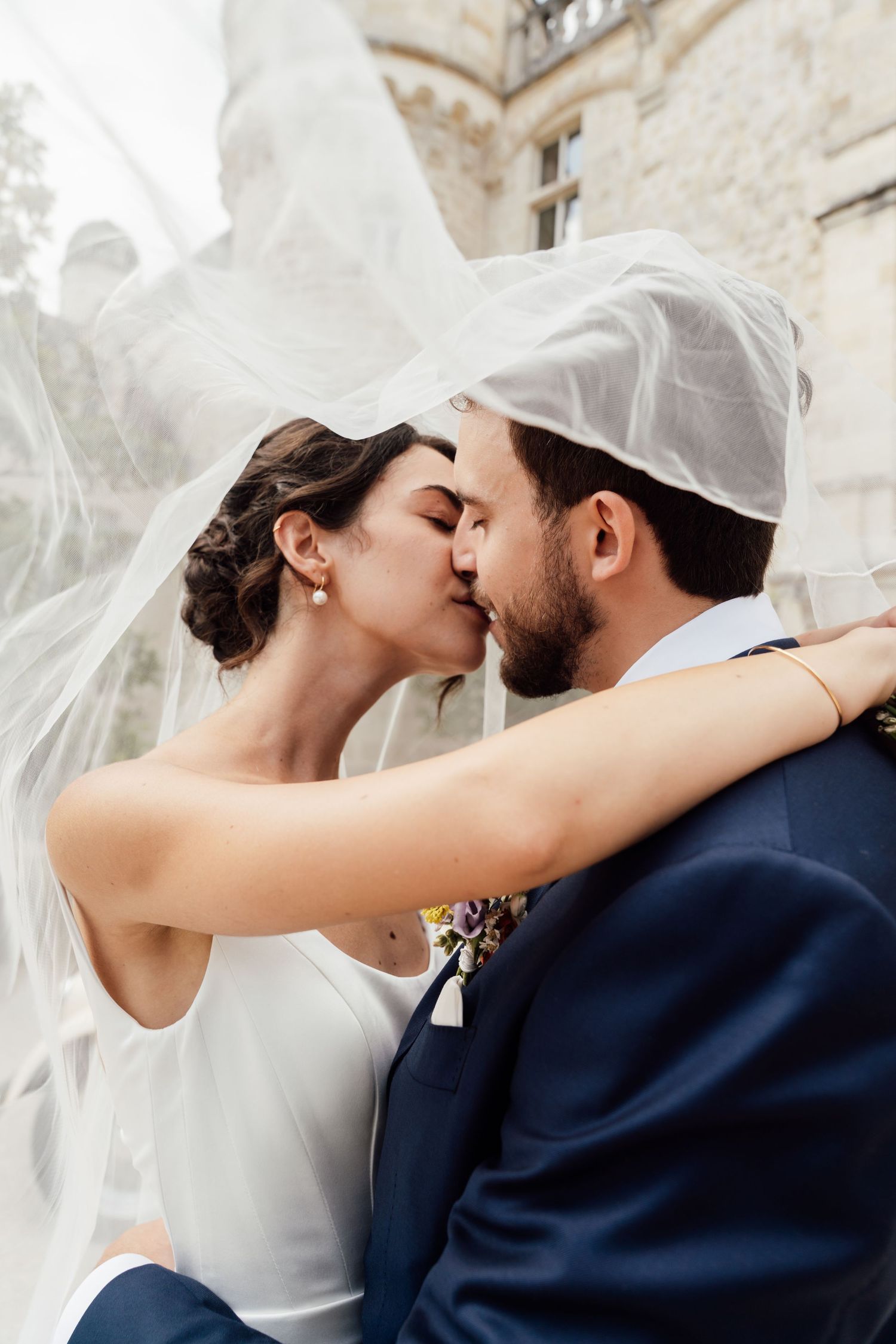 Bride and groom sharing a romantic kiss under a flowing white veil outdoors near a stone building.