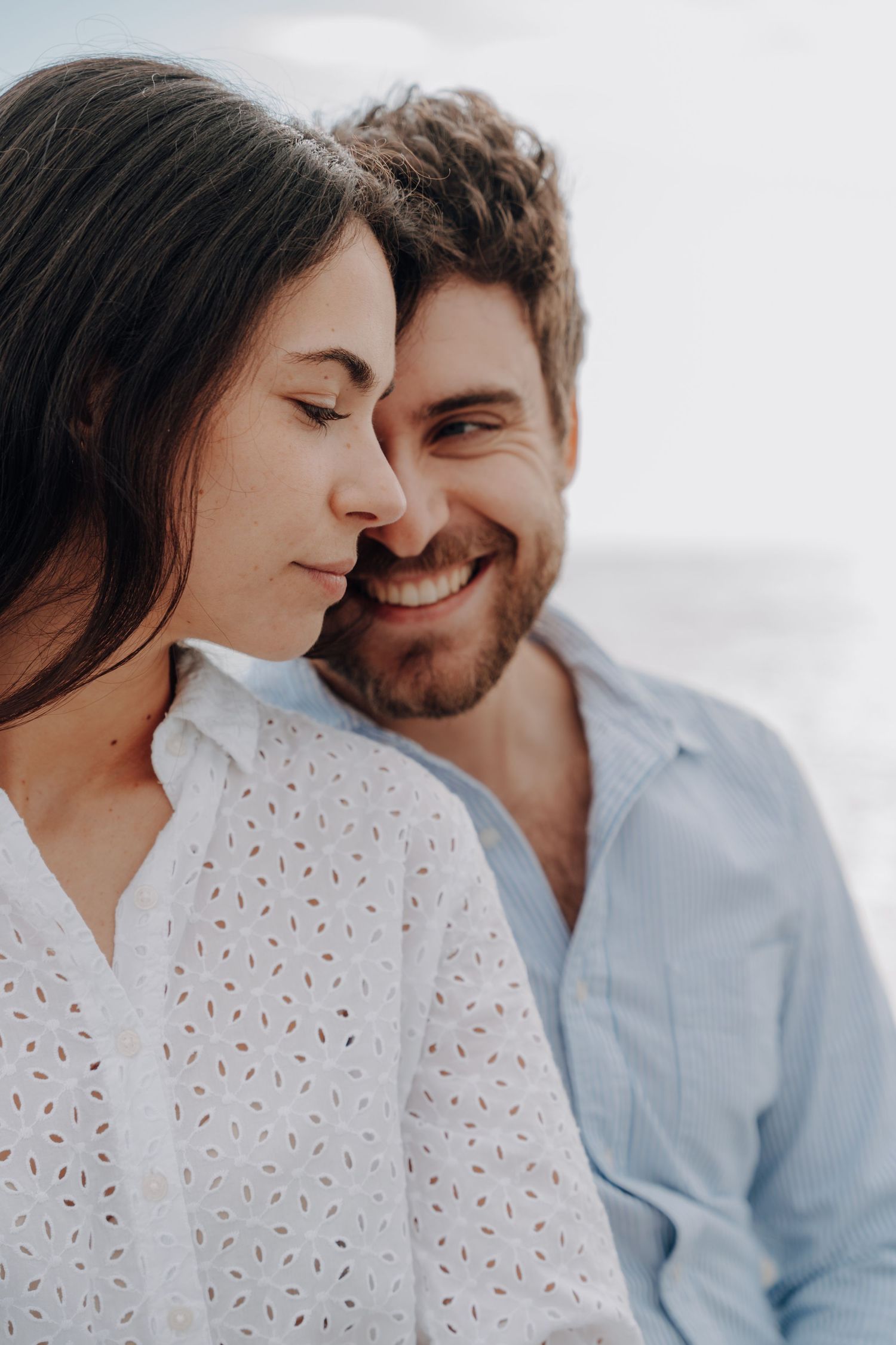 A couple in blue and white outfits shares an intimate moment during a beach photoshoot.
