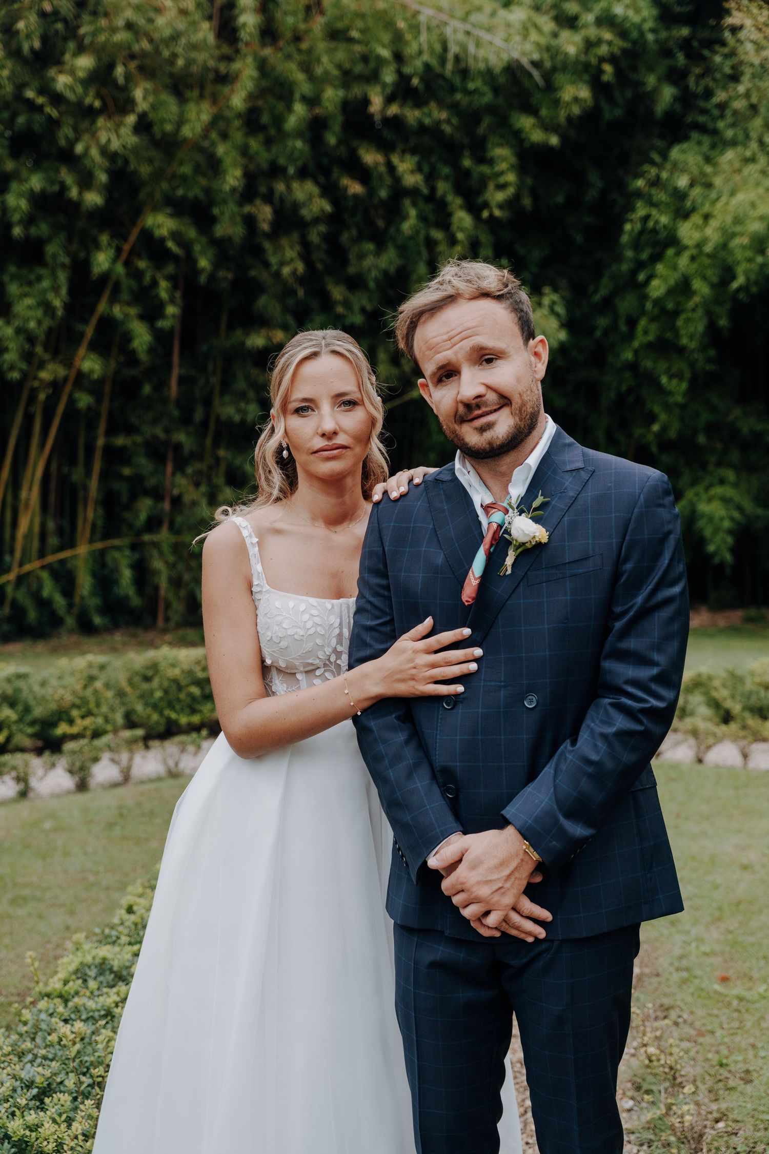Bride and groom pose together in formal wedding attire in garden setting.