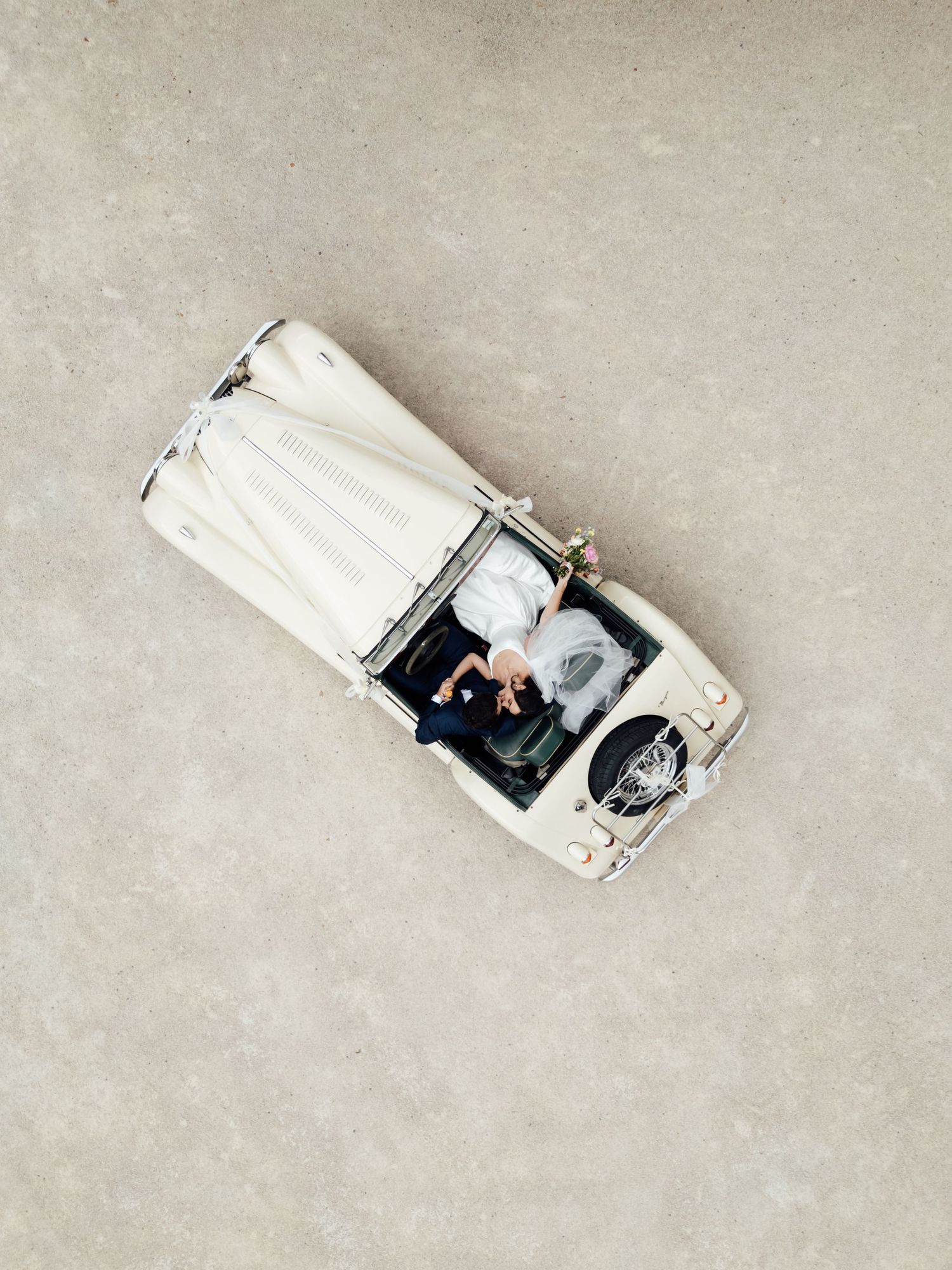 Aerial view of a couple in a vintage white convertible car photographed from above on a neutral surface.