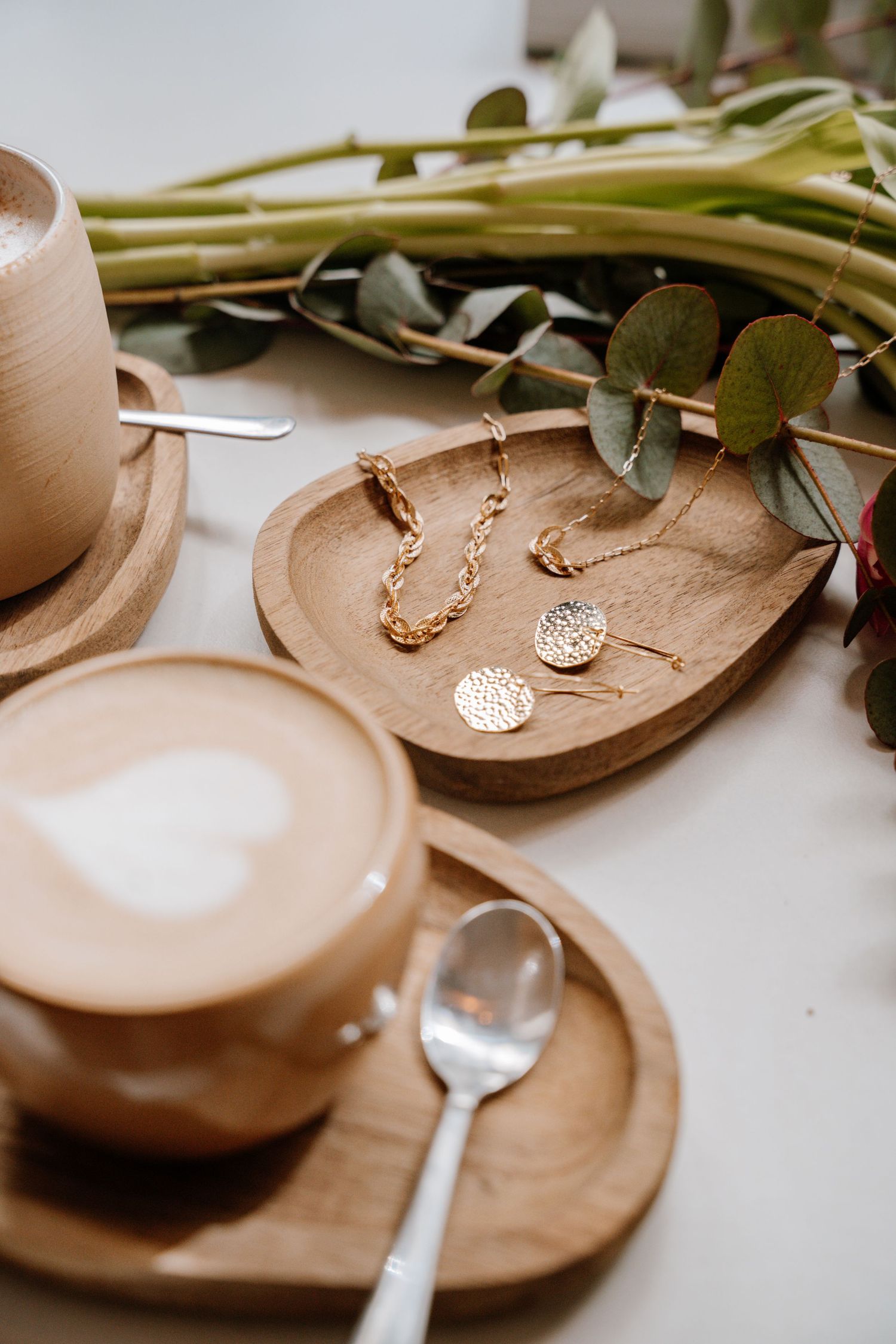 Gold pendant necklaces with hammered disc charms displayed on wooden plates with eucalyptus stems and white candles.